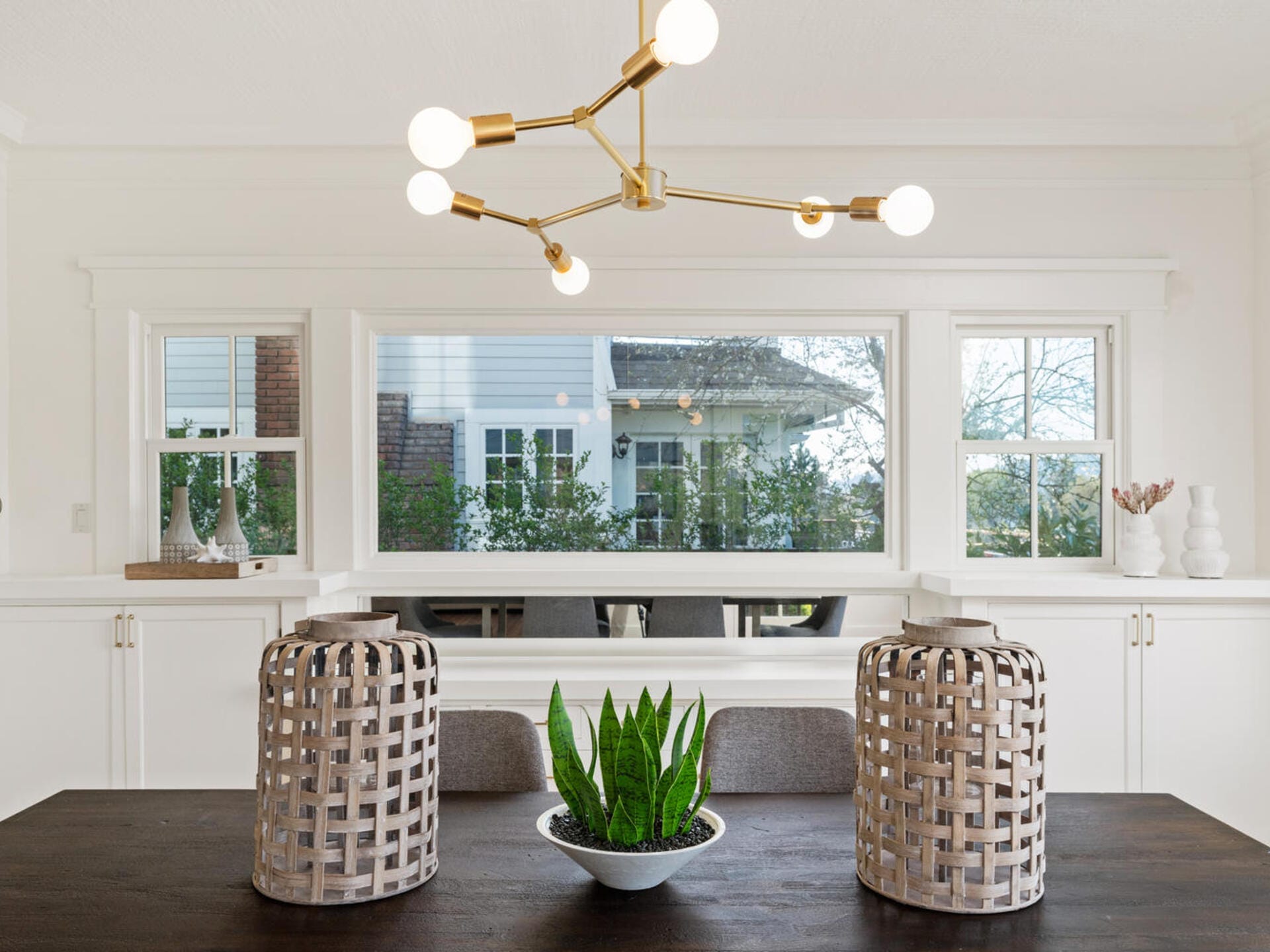 A modern dining room features a dark wood table with a potted plant centerpiece and two large wicker lanterns. A brass chandelier with globe lights hangs above. Large windows offer a view of greenery and a white house exterior.