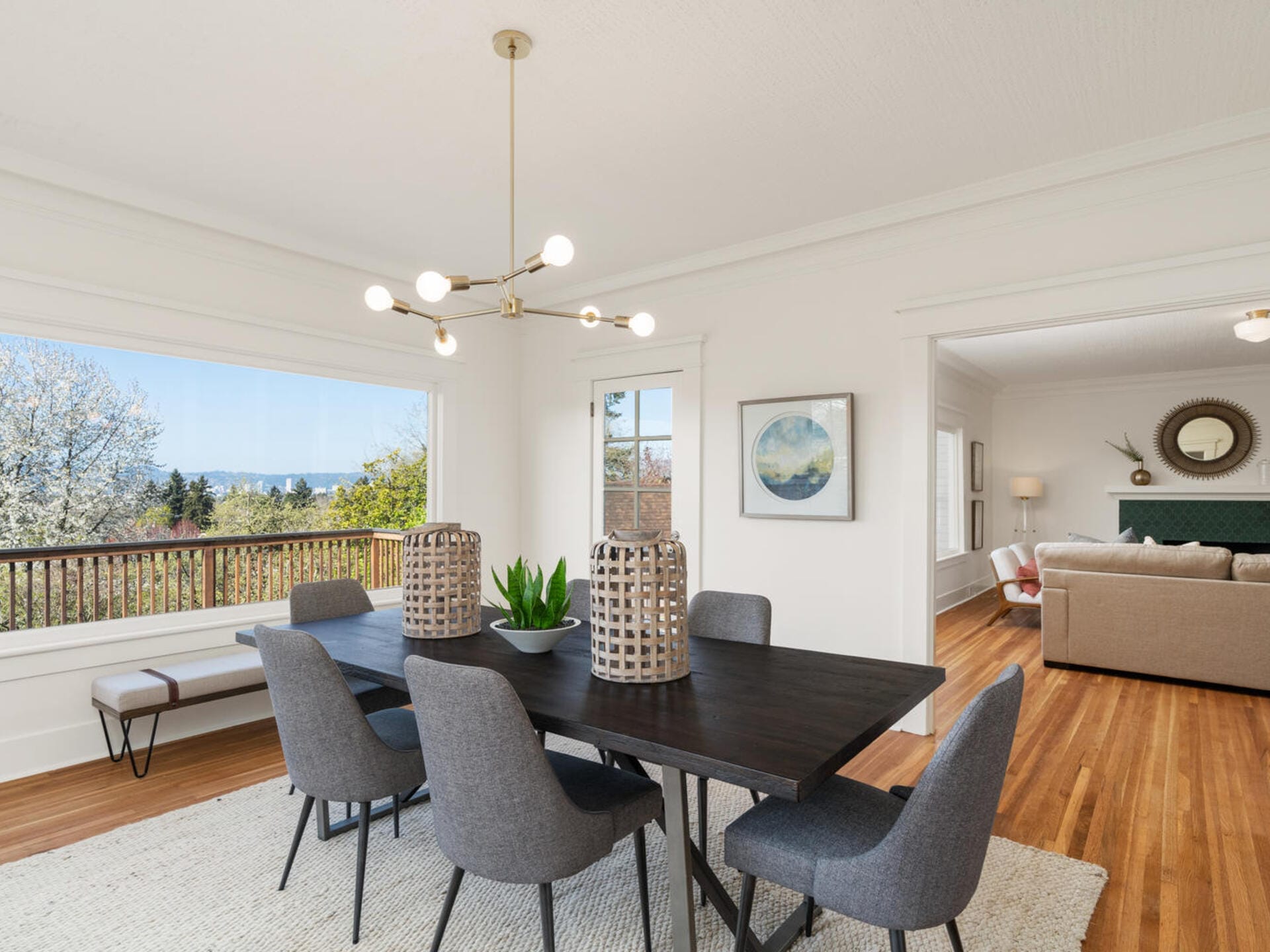 A bright dining room with a dark wooden table and six gray chairs. A modern chandelier hangs above the table, which is decorated with a plant. Large windows showcase a scenic outdoor view. Nearby, a living room features a sofa and decorative items.