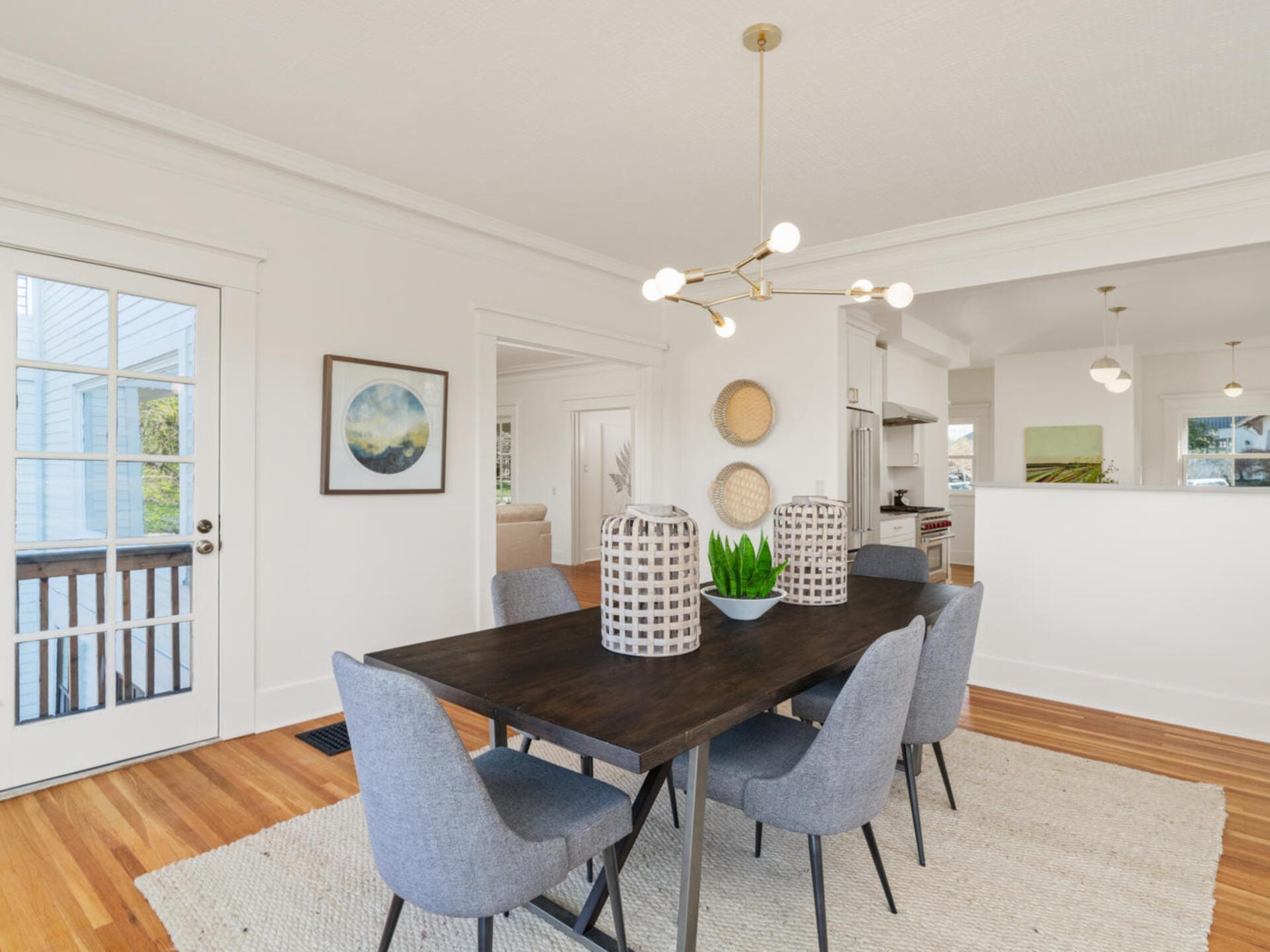 A modern dining room with a dark wooden table and six gray upholstered chairs. A contemporary light fixture hangs above. The room is bright, with white walls, a large rug, and decorative elements, including a potted plant and wall art.