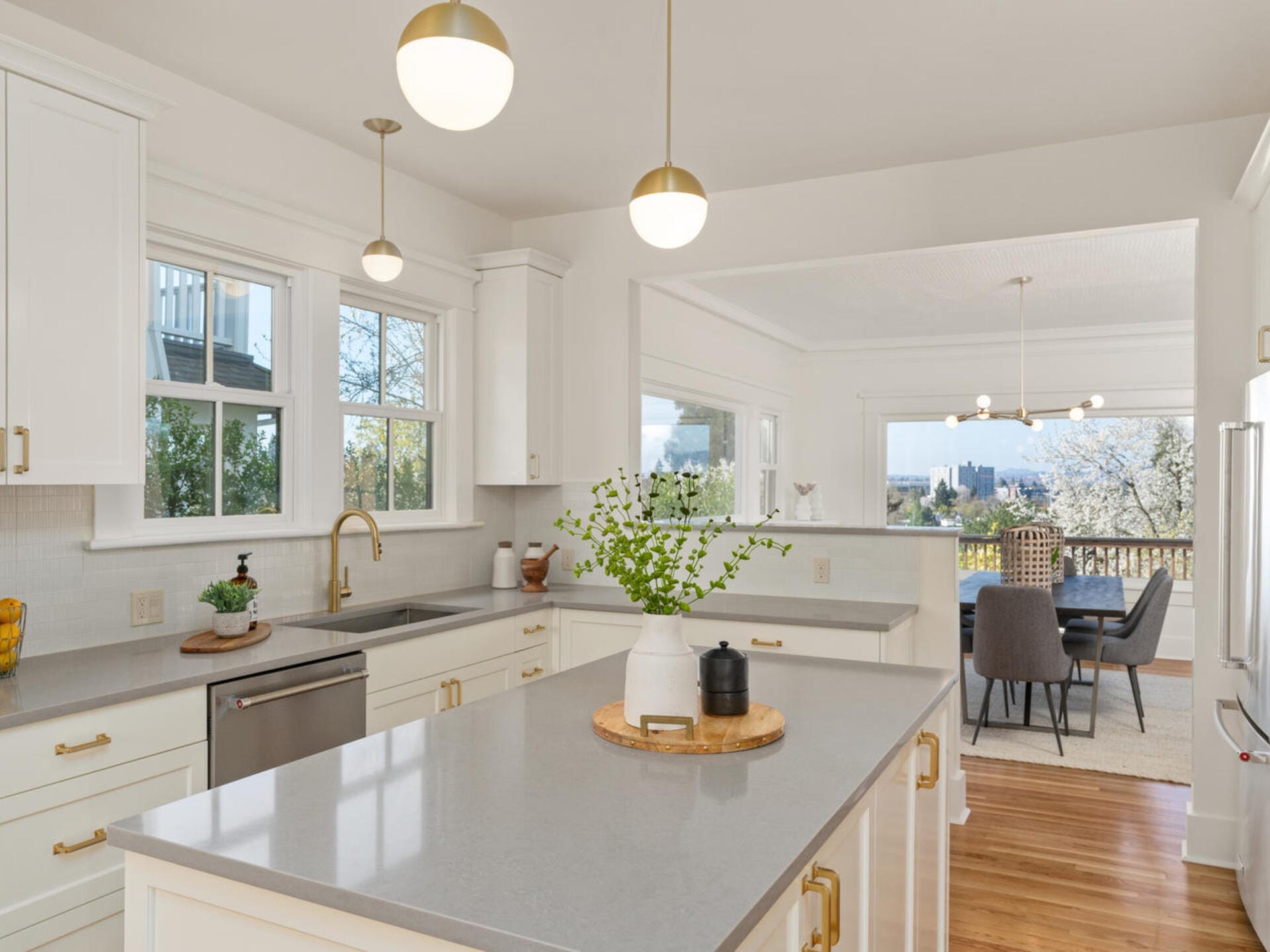 A bright, modern kitchen with light gray countertops, white cabinets, and gold handles. Pendant lights hang above the island, which features a vase with greenery. In the background, a dining area with large windows offers a scenic view.