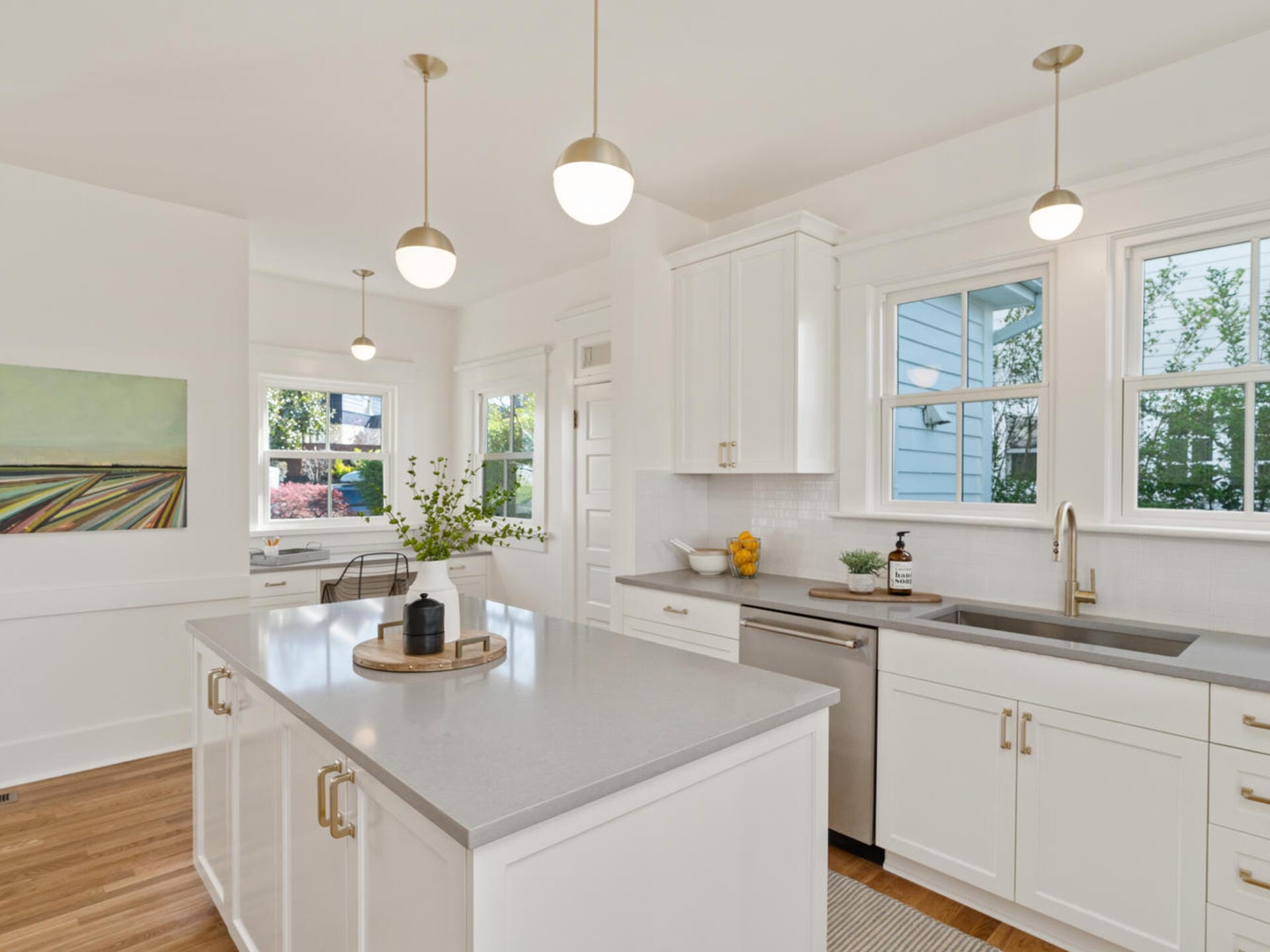 A modern kitchen with white cabinetry and a large island featuring a gray countertop. Three pendant lights hang above. Natural light streams in from multiple windows, highlighting a vase with greenery and a small bowl of lemons on the counter.