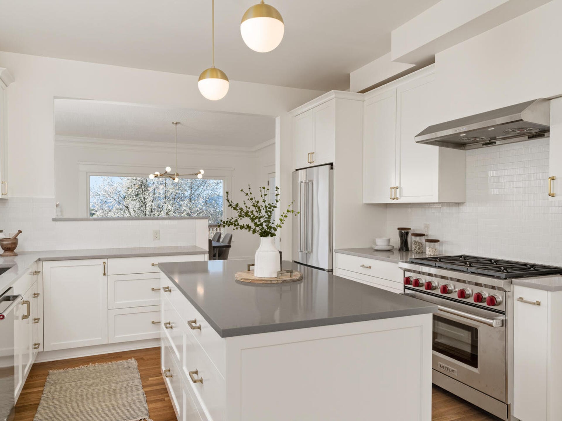 A modern kitchen with white cabinetry, stainless steel appliances, and a gray countertop island. Two pendant lights hang above the island. A vase with greenery sits on the island. A large window provides natural light and a view of trees.