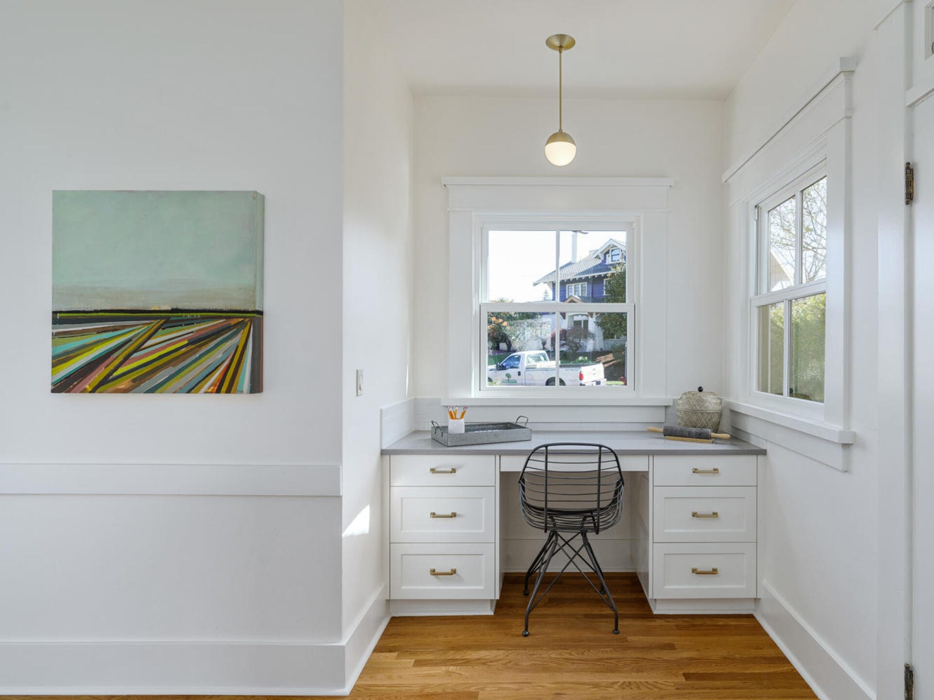A bright home office with a white desk beneath a window, featuring a wire chair and a laptop. A colorful abstract painting hangs on the left wall. The room has wooden floors and a single ceiling light fixture.