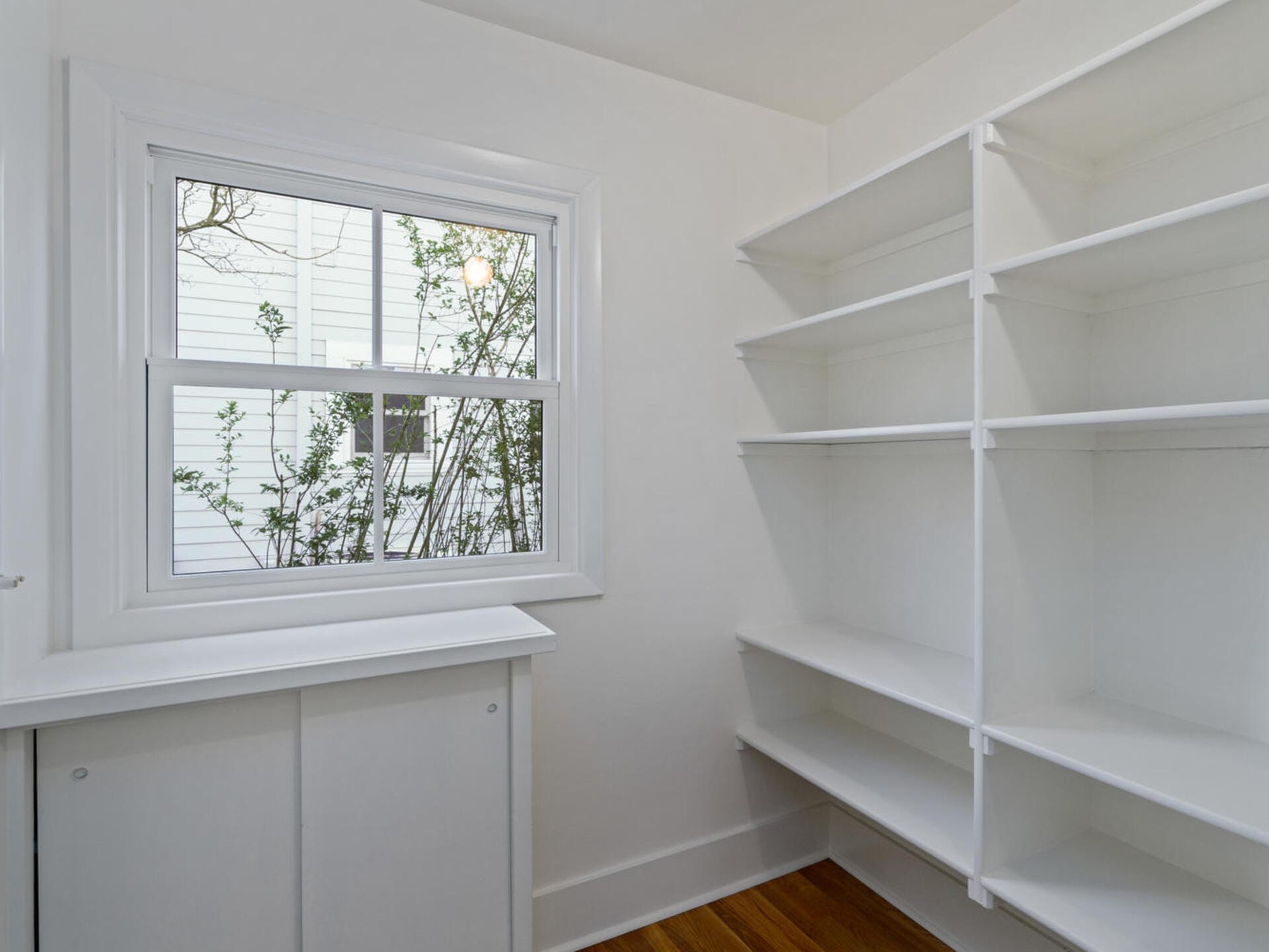 A small room with white walls features built-in shelving on one side and a window on the opposite wall. The floor is wooden, and the shelves are empty, creating a clean and minimalist appearance.