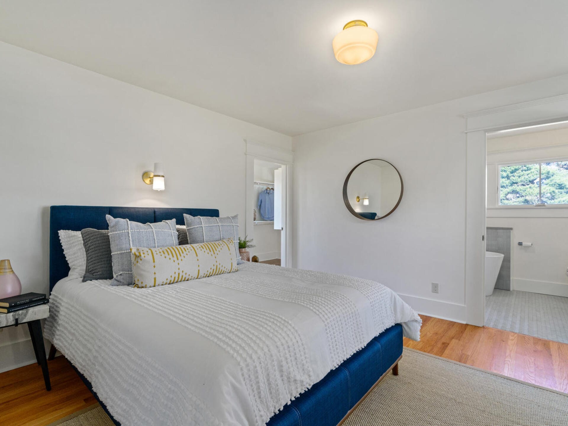 A cozy bedroom with a dark blue bed featuring white and patterned pillows. The room has white walls, a round mirror, and wooden floors. A doorway opens to a bright bathroom with a visible bathtub. Soft overhead lighting illuminates the space.