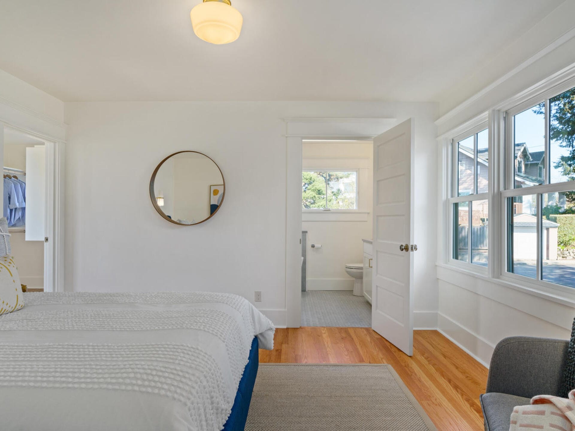 A bright bedroom featuring a neatly made bed with a textured white bedspread. A round mirror hangs on the wall, and a door opens to a bathroom. Large windows on the right let in natural light. A small chair is visible in the corner.