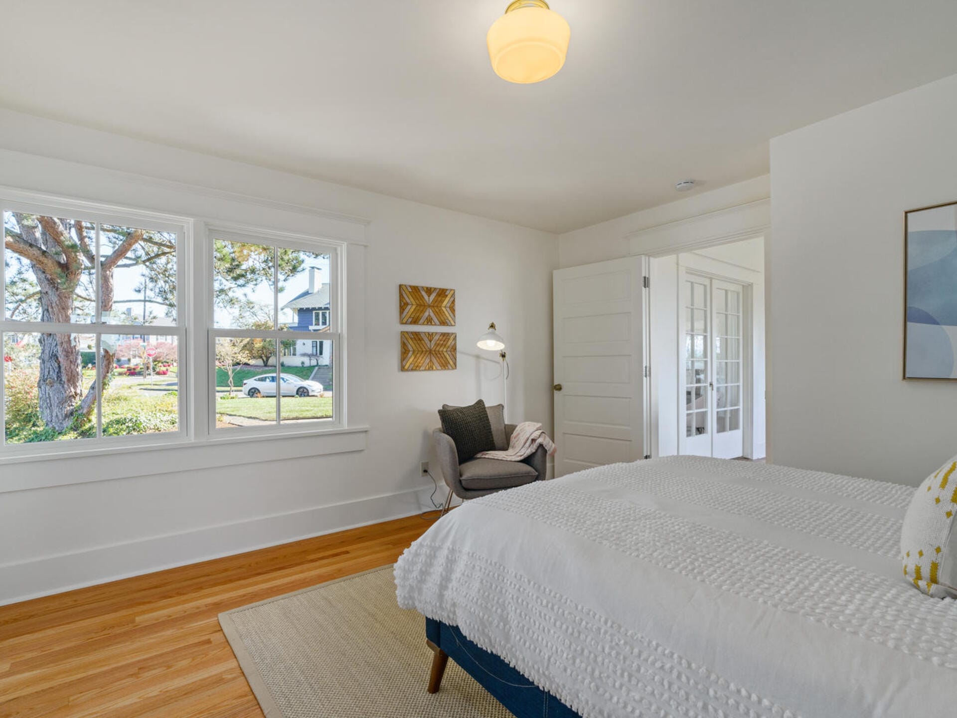 A bright bedroom with a large window showing a tree and a house outside. The room features a bed with a white bedspread, a cozy armchair with a lamp beside it, and wall art. The floor is wooden with a beige rug.