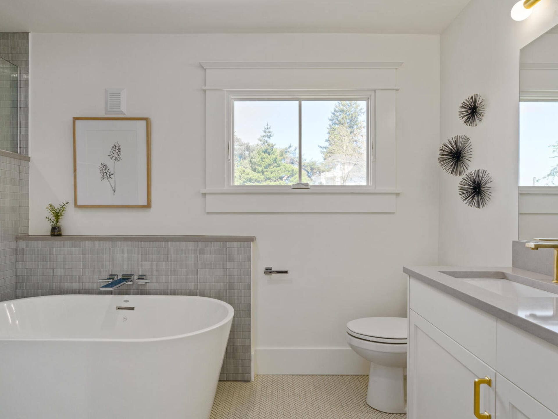A modern bathroom with a freestanding bathtub, a wall-mounted toilet, and a minimalist vanity. The walls are painted white and feature abstract decorations. A window above the bathtub lets in natural light.
