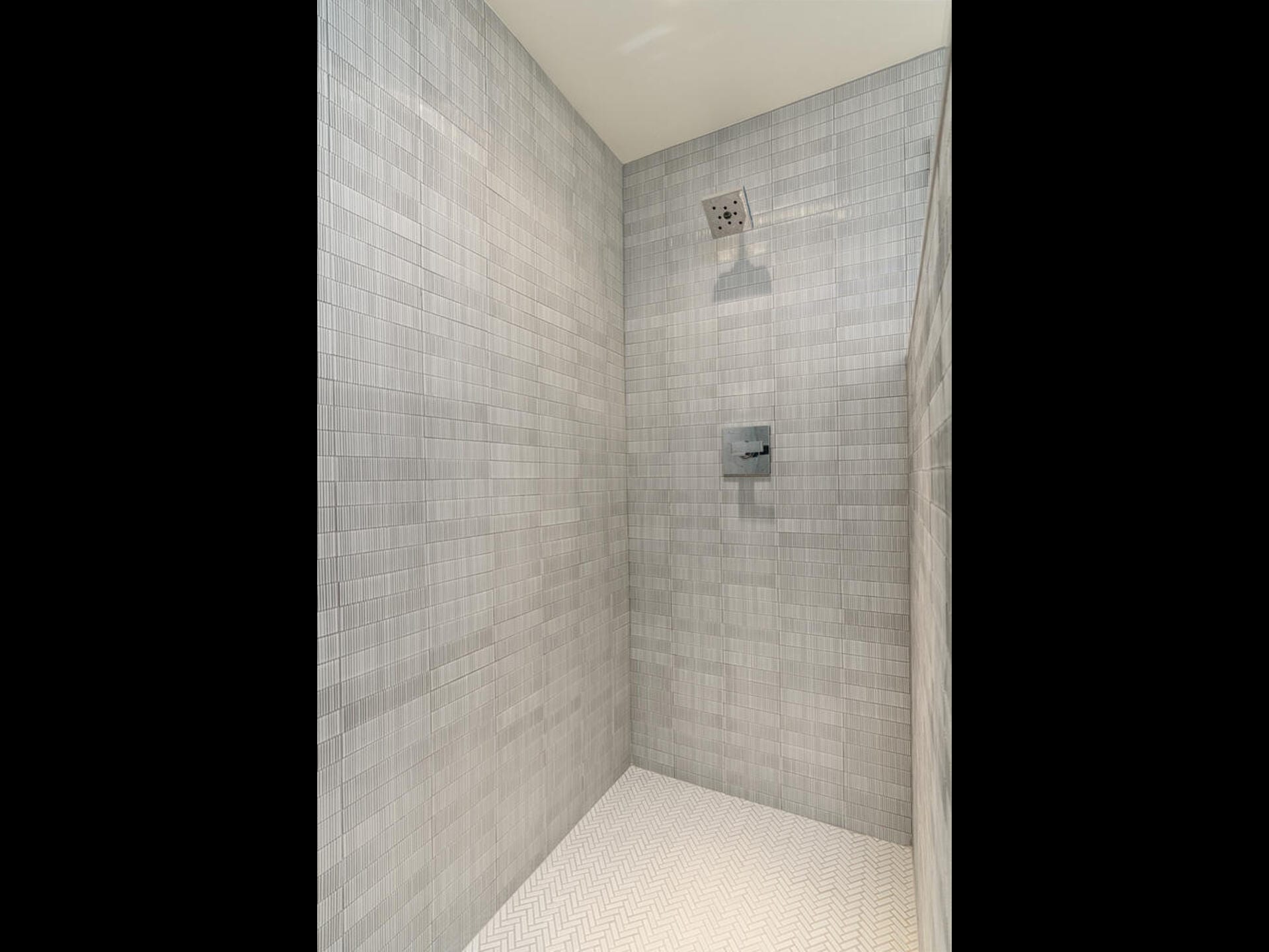 A modern shower with light gray tile walls and a patterned white tile floor. It features a square rainfall showerhead and a small wall-mounted control panel.