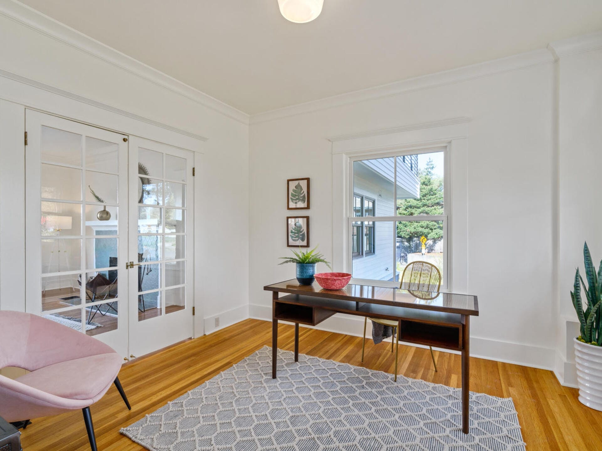 A bright home office with a wooden desk and chair on a gray patterned rug. The room features a pink armchair, potted plants, wall art, and a large window. French doors open to another room with more seating and decor. Natural light fills the space.
