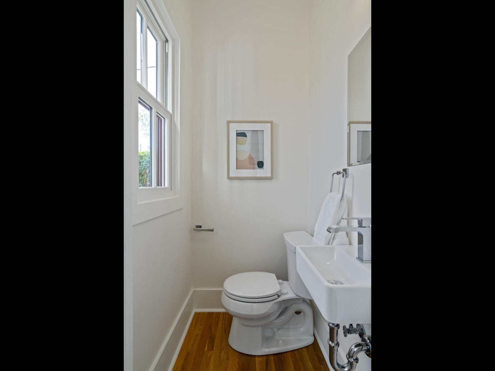 A small, modern bathroom with white walls features a window, abstract art, toilet, and wall-mounted sink with a hand towel. The room has hardwood flooring and minimalistic decor.