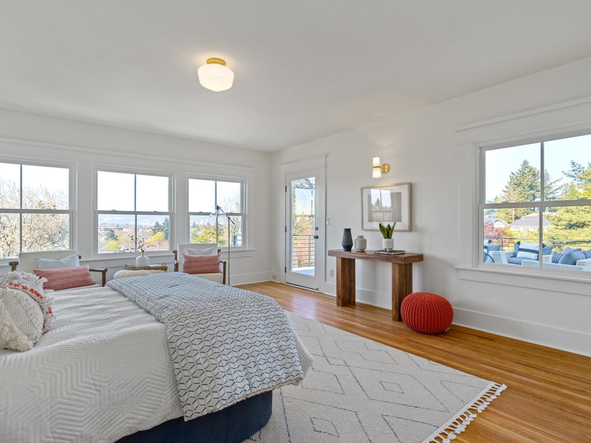 A bright bedroom with large windows, a bed with white linens and patterned pillows, a wooden floor, and a rug. Theres a wooden dresser with decor, a red pouf, and a door leading to a balcony with views of greenery outside.