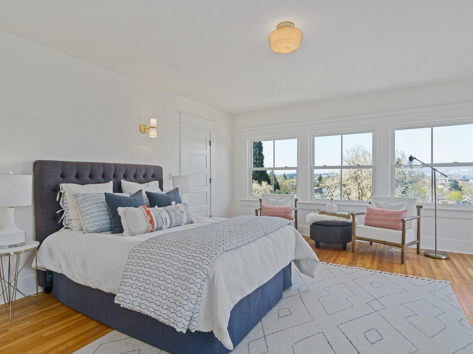 A bright bedroom with a large gray upholstered bed adorned with multiple pillows and a patterned blanket. The room features a window with a scenic view, a cozy seating area with armchairs, and a white rug on wooden floors.