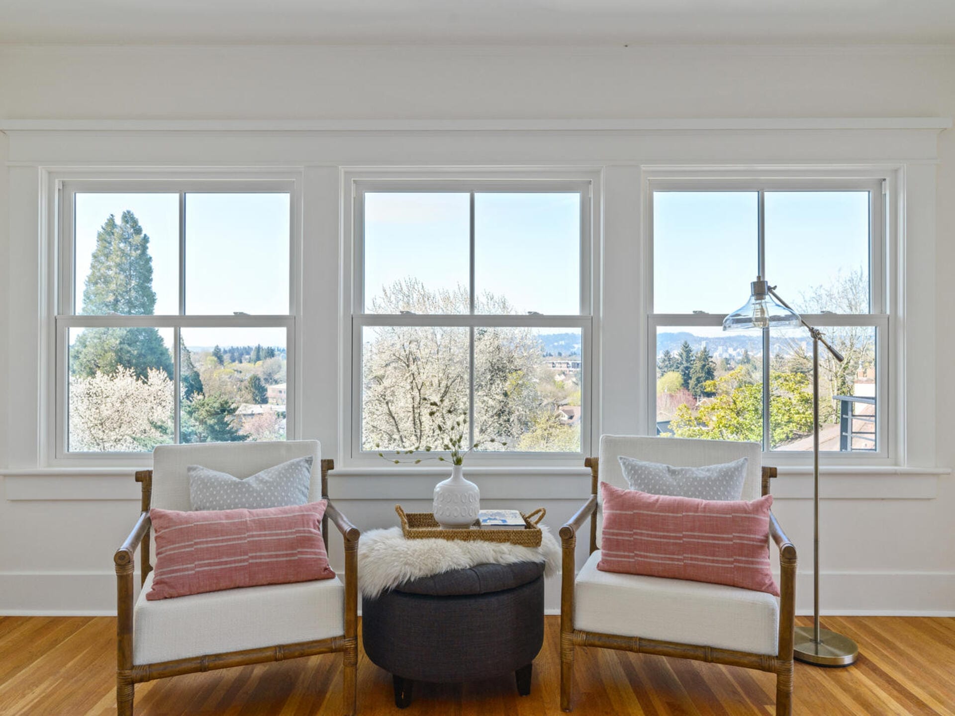 A bright room with three large windows showing a scenic view. Two armchairs with pink pillows flank a small gray ottoman, topped with a vase on a tray. A standing lamp is beside the chairs, and trees are visible outside.