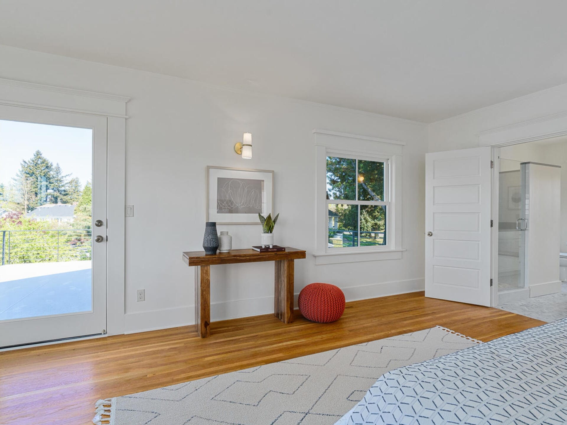 A bright bedroom with a wooden floor, white walls, and a rug. Theres a bed on the right, a door leading to a patio, a wooden table with decor items, and a red pouf on the floor. A bathroom entrance is visible on the left.