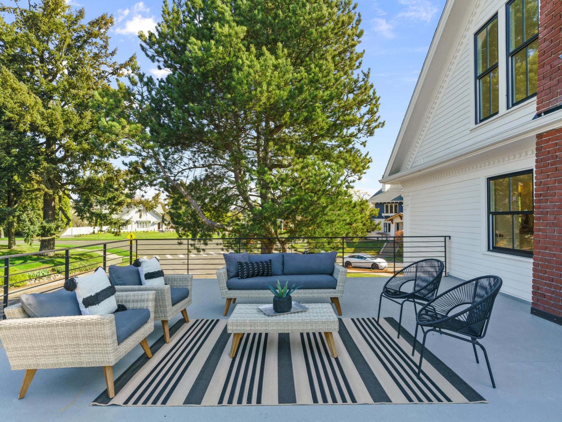 Outdoor patio with cushioned wicker sofas and chairs arranged on a striped rug. A small coffee table with a plant sits in the center. The area is surrounded by trees and faces a two-story white house with a brick accent wall.