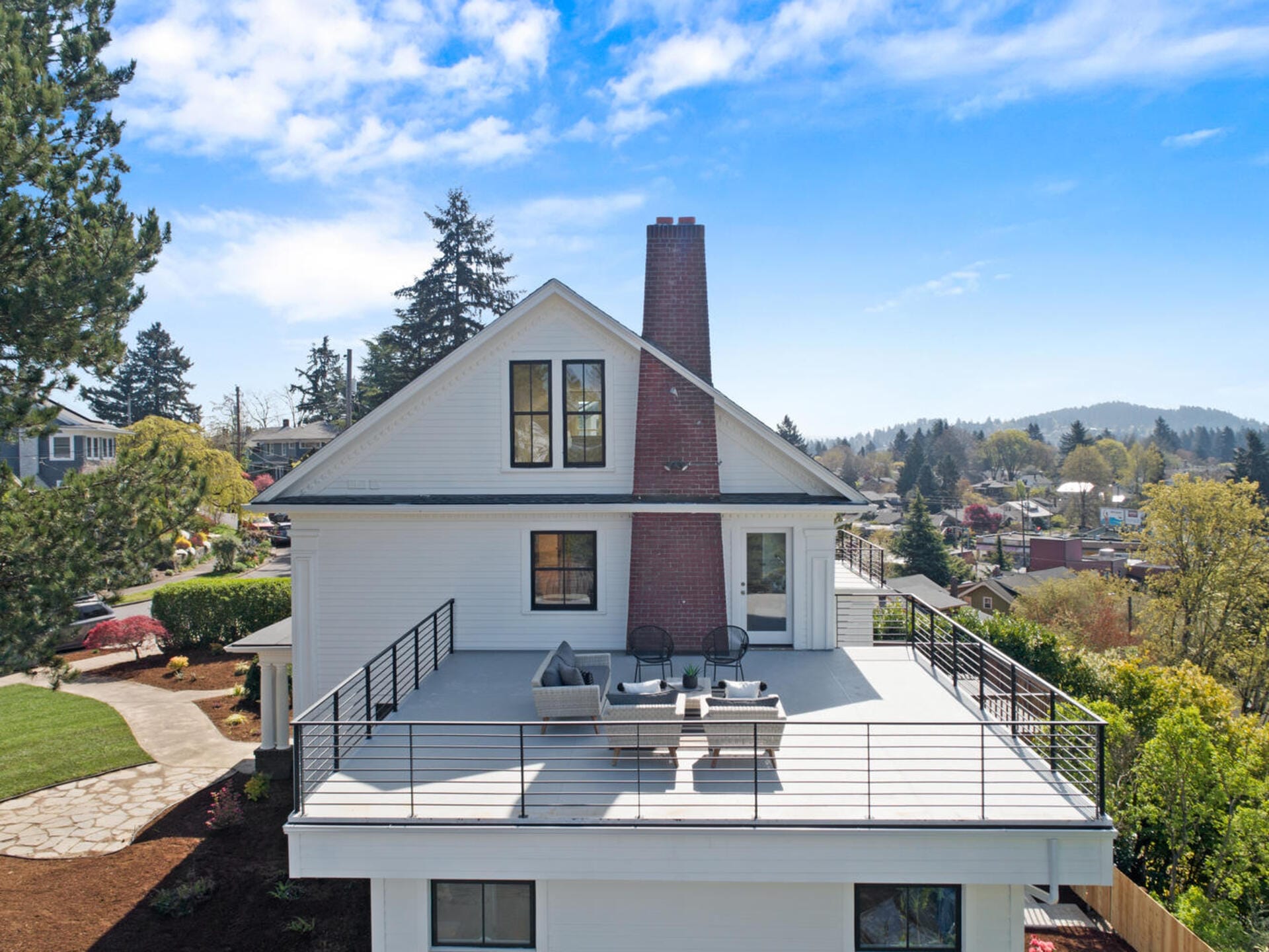 A white two-story house with a large rooftop deck featuring outdoor furniture. A red brick chimney extends from the deck. The house is surrounded by green trees and a neighborhood, with a clear blue sky overhead.