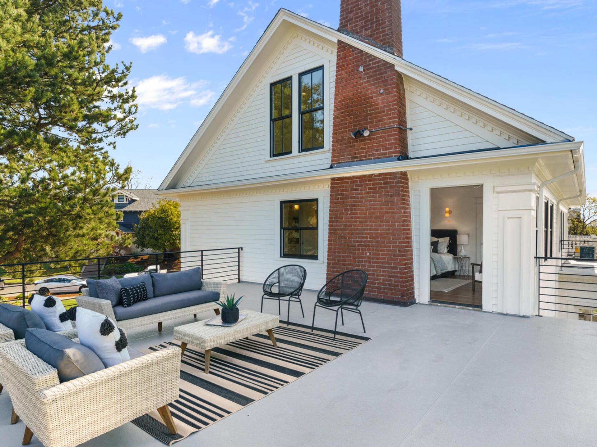 A spacious rooftop patio features wicker sofas, black chairs, and a striped rug, set against a white house with a prominent brick chimney. A bedroom with a neatly made bed is visible through an open door. Trees and a clear blue sky are in the background.