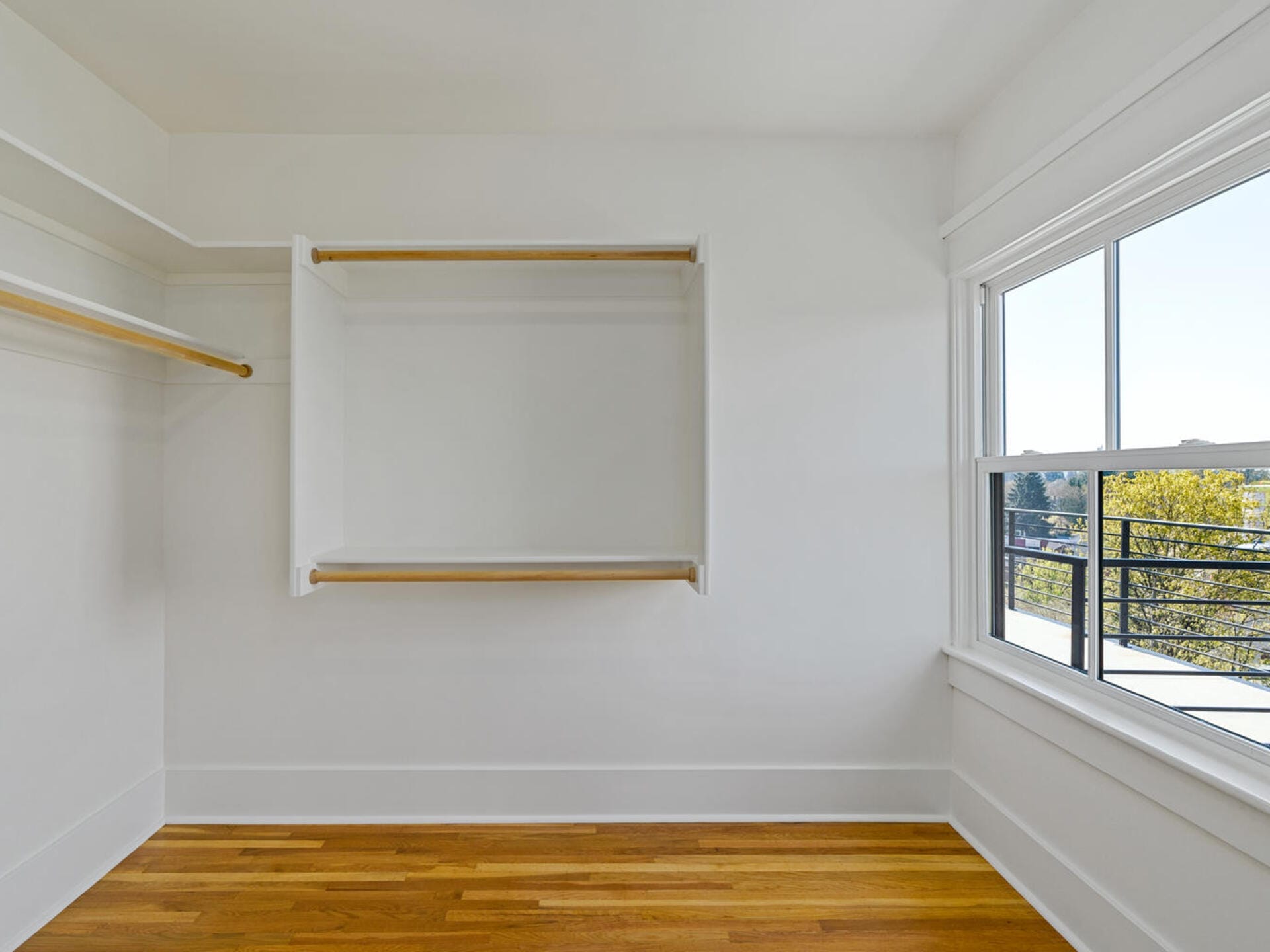 An empty walk-in closet with wooden floors and white walls. It features two wooden rods for hanging clothes and a large window on the right side, offering a view of a balcony and trees outside.