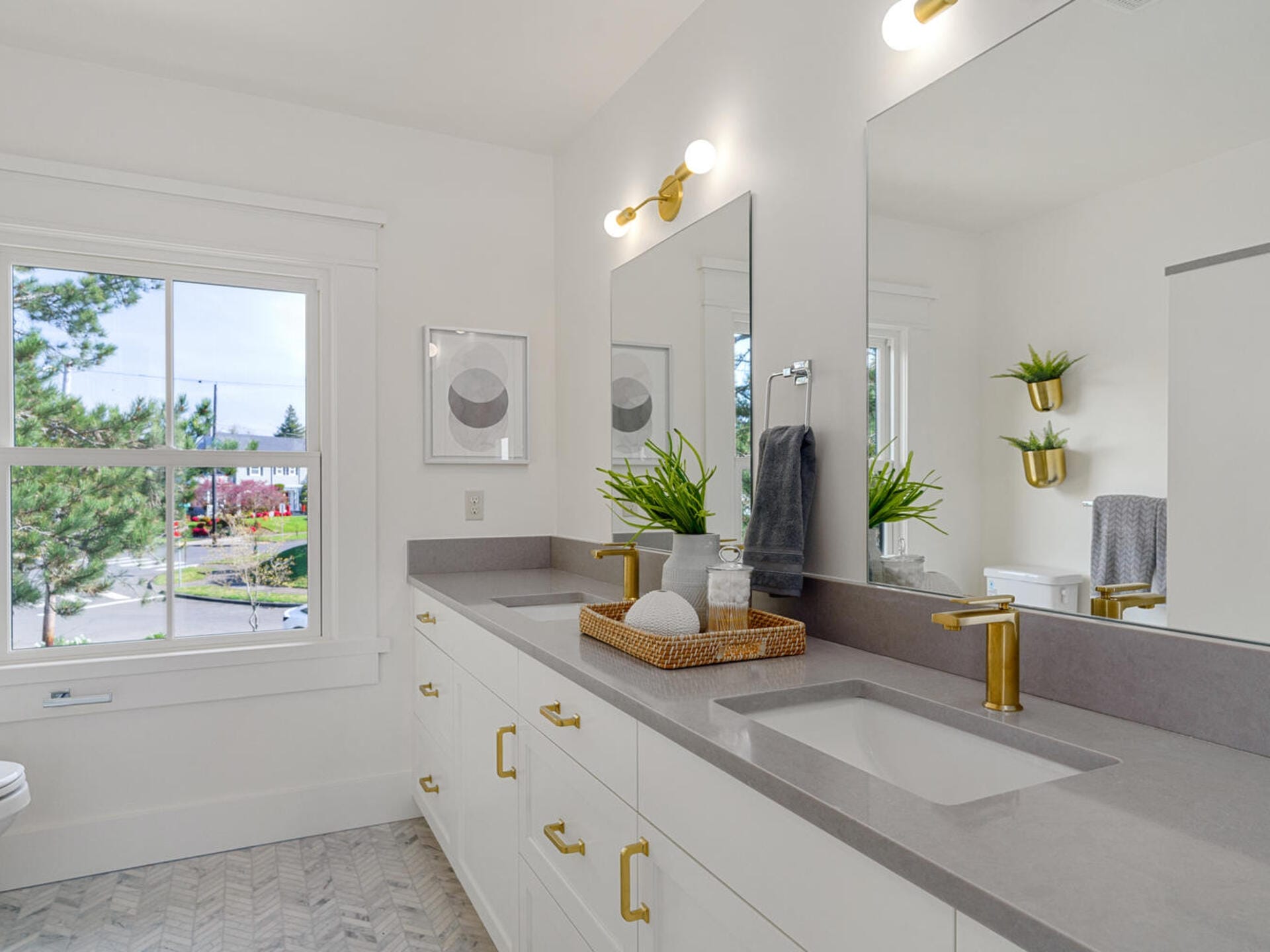 Modern bathroom with a double vanity, gray countertop, and gold fixtures. Two sinks, two mirrors, potted plants, and wall art are visible. A window shows a view of trees and a neighborhood. The floor has a herringbone pattern.
