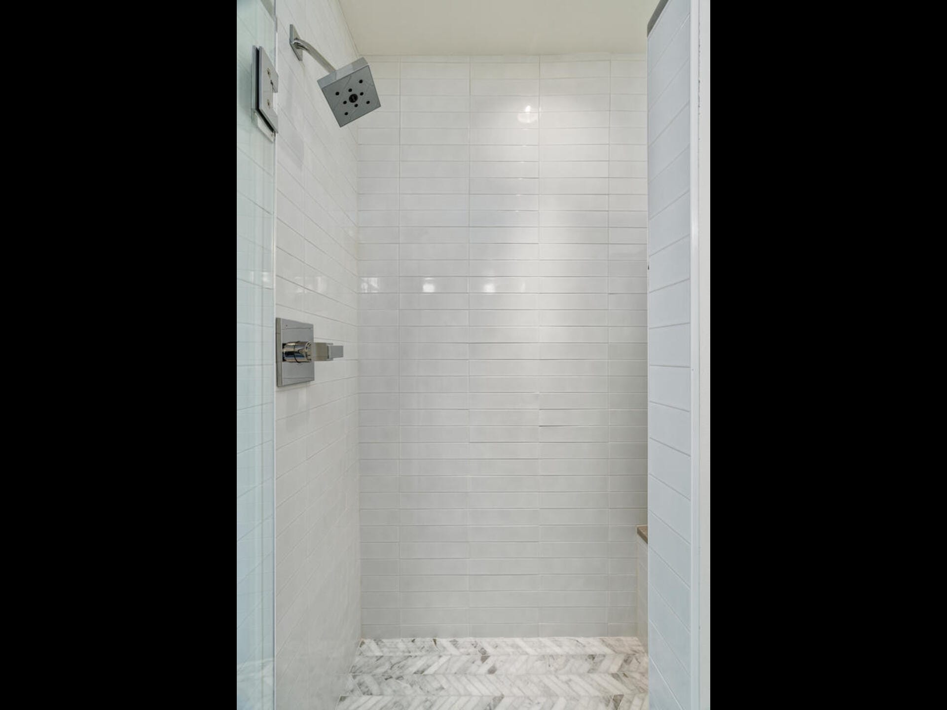 A modern shower with white subway tiles, a square rainfall showerhead, and a sleek silver control panel. The floor is covered in a herringbone-patterned marble tile. The lighting is soft and evenly distributed.