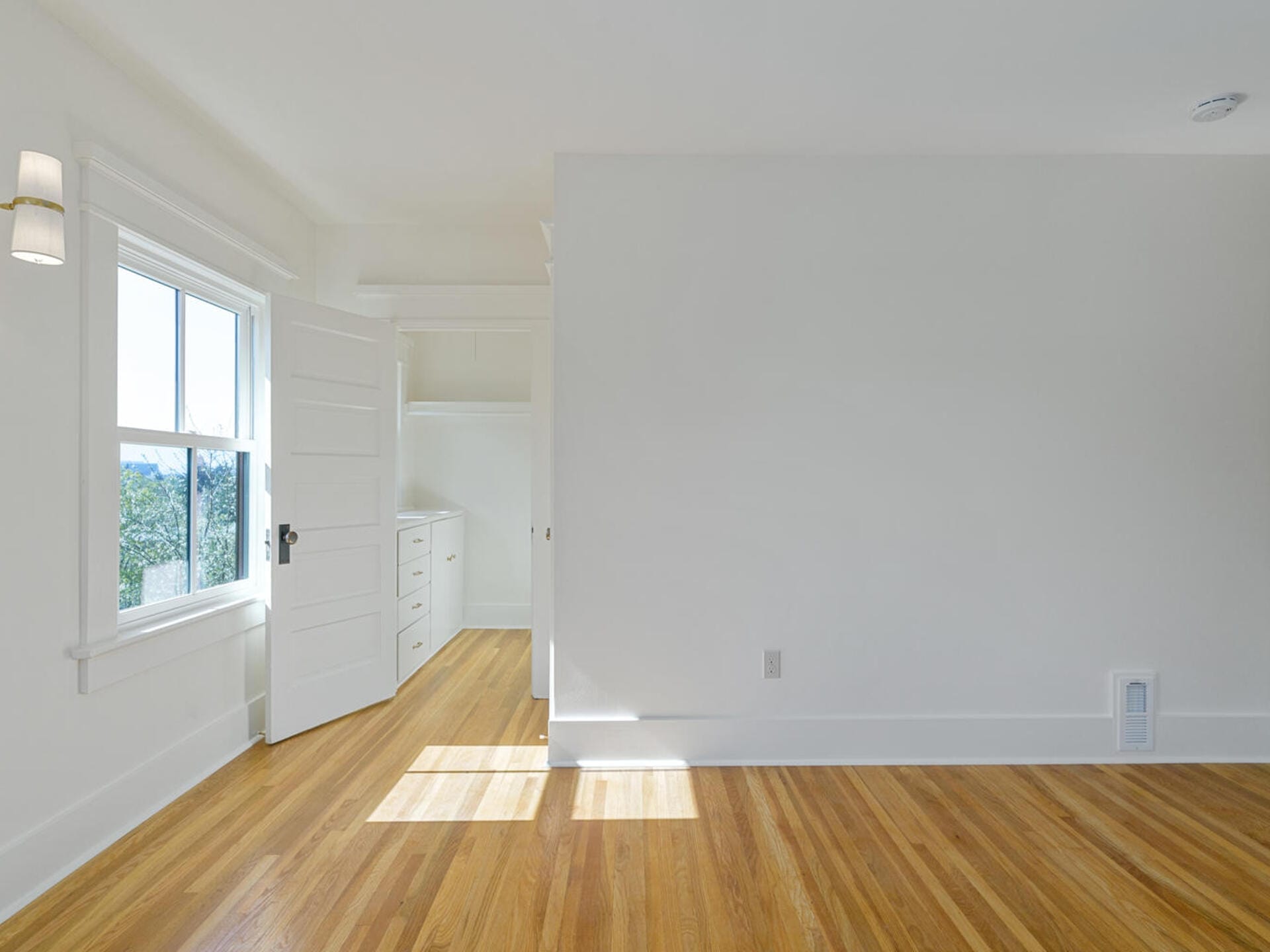 A bright, empty room with light wooden floors and white walls. Sunlight streams through a single window on the left. An open door leads to a small alcove with built-in drawers, and a modern wall sconce is on the left wall.