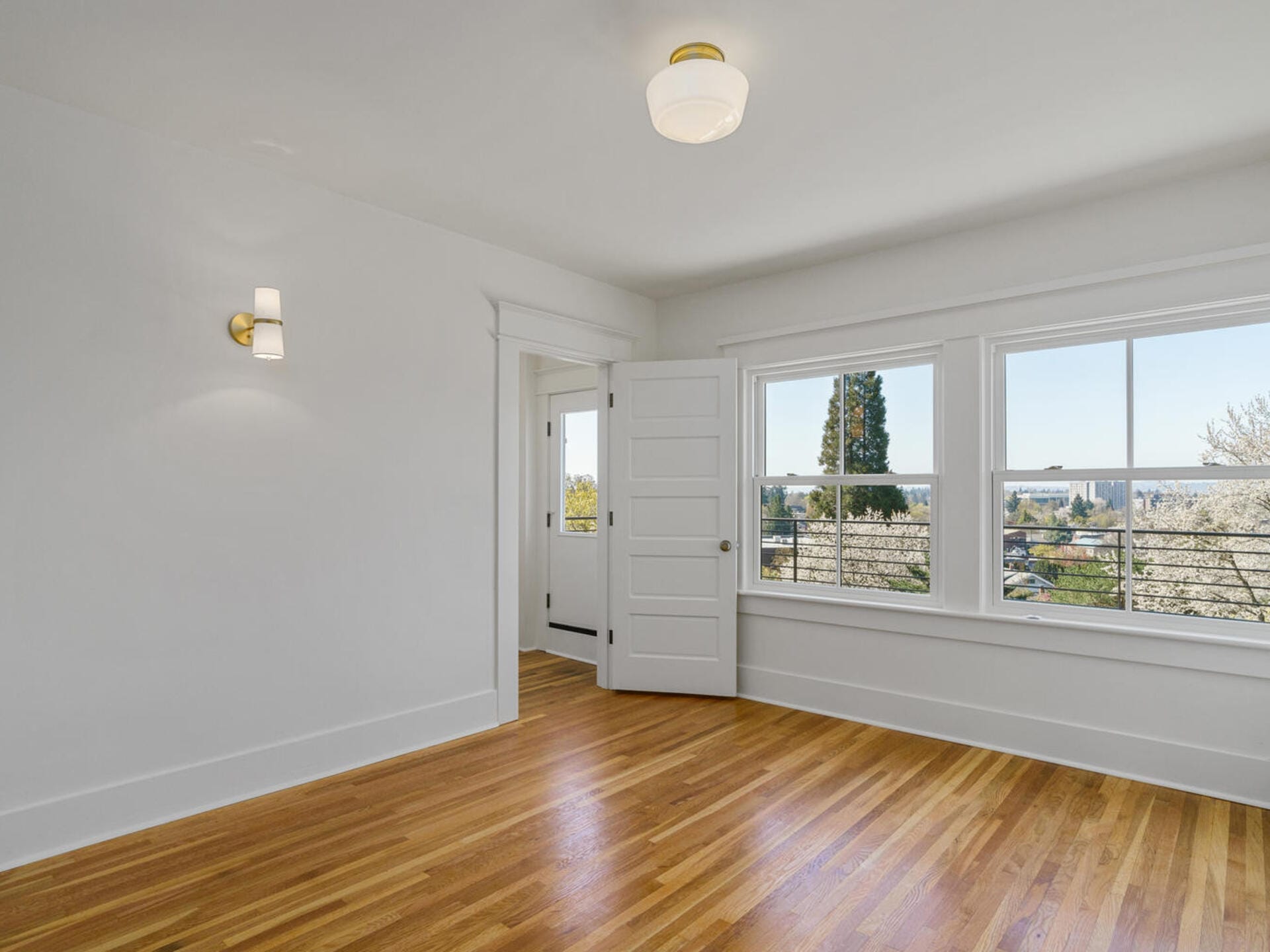 An empty room with light wooden flooring, white walls, and a ceiling light. There are two large windows with a view of trees and a partially open door leading to another room. Natural light fills the space.