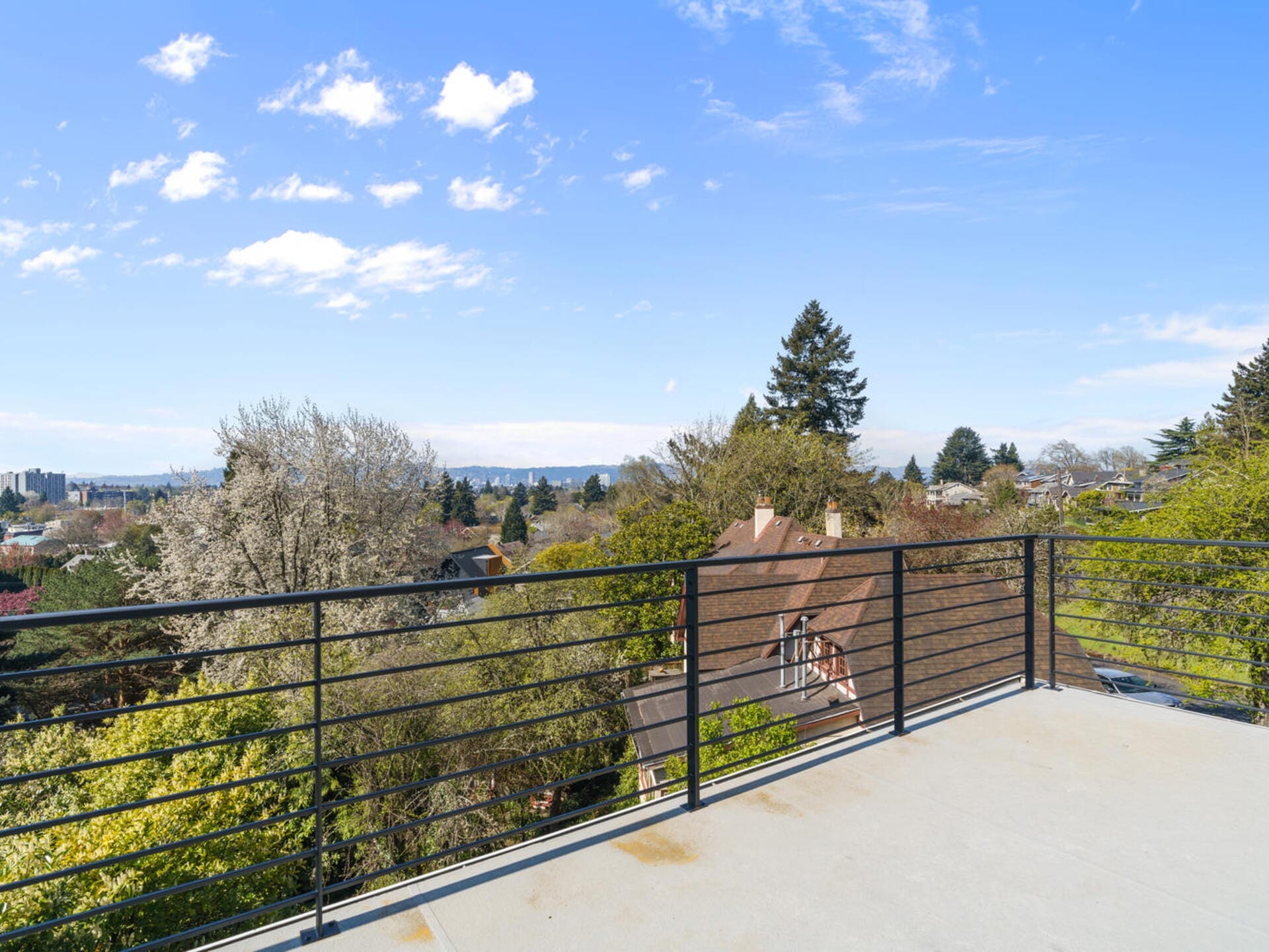 View from a balcony with a black railing overlooking a neighborhood with trees and houses. The sky is clear with scattered clouds, and a city skyline is visible in the distance.