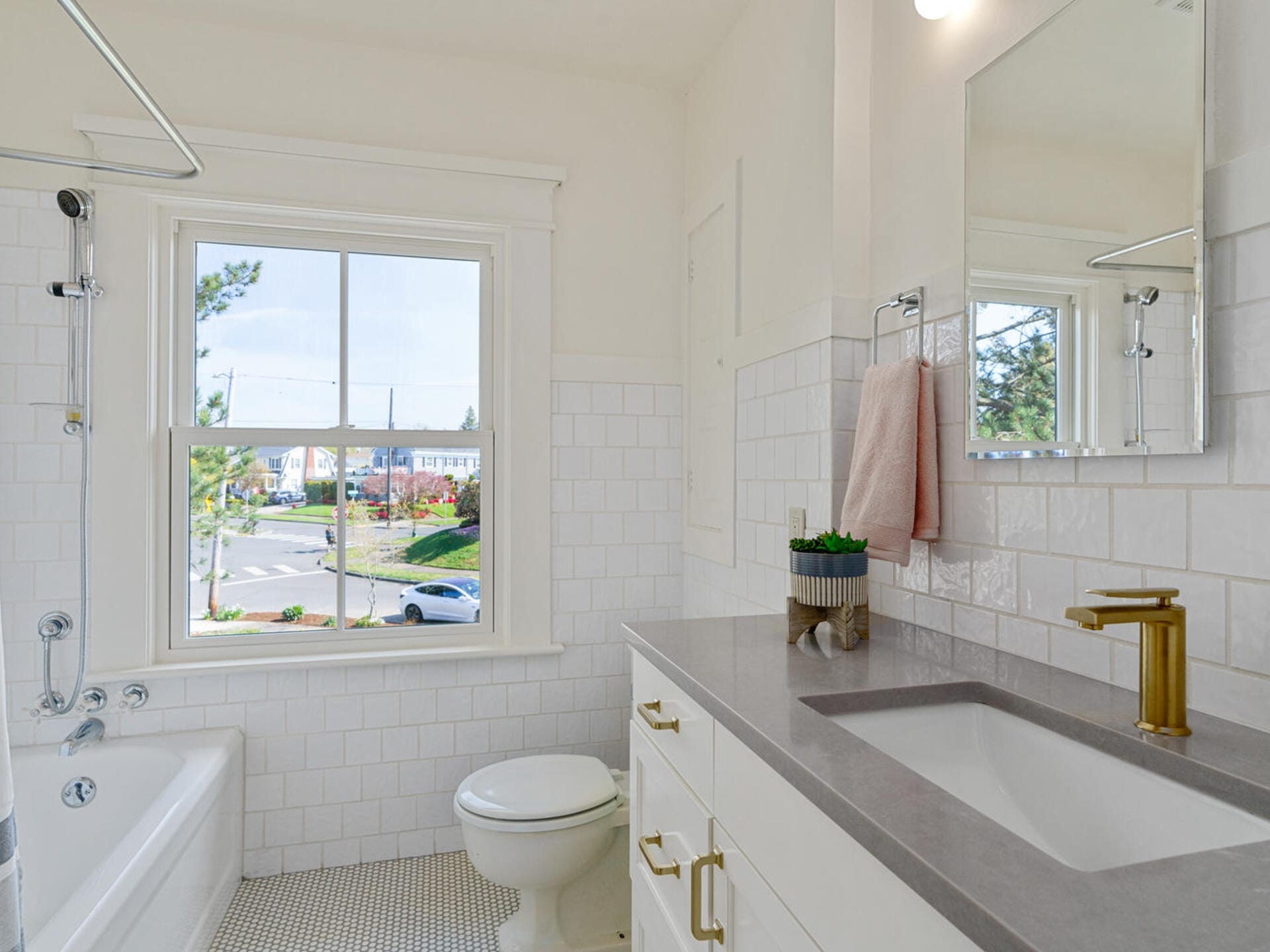 A bright bathroom with white tiled walls, a bathtub, toilet, and a gray countertop sink. Gold faucet and handles accent the cabinetry. A window shows a view of a street and trees outside. A pink towel and a small plant are near the sink.