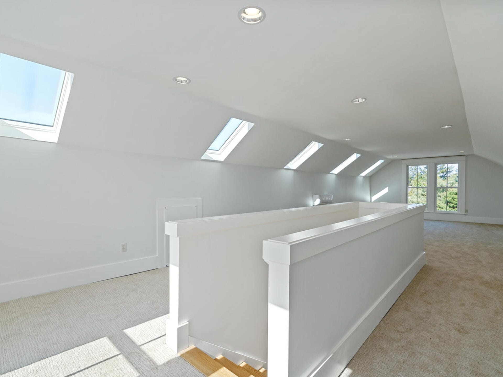 A spacious white attic room with multiple skylights, beige carpet, and recessed lighting. A long white railing runs the length of the room, leading to double glass doors at the far end, allowing natural light to fill the space.