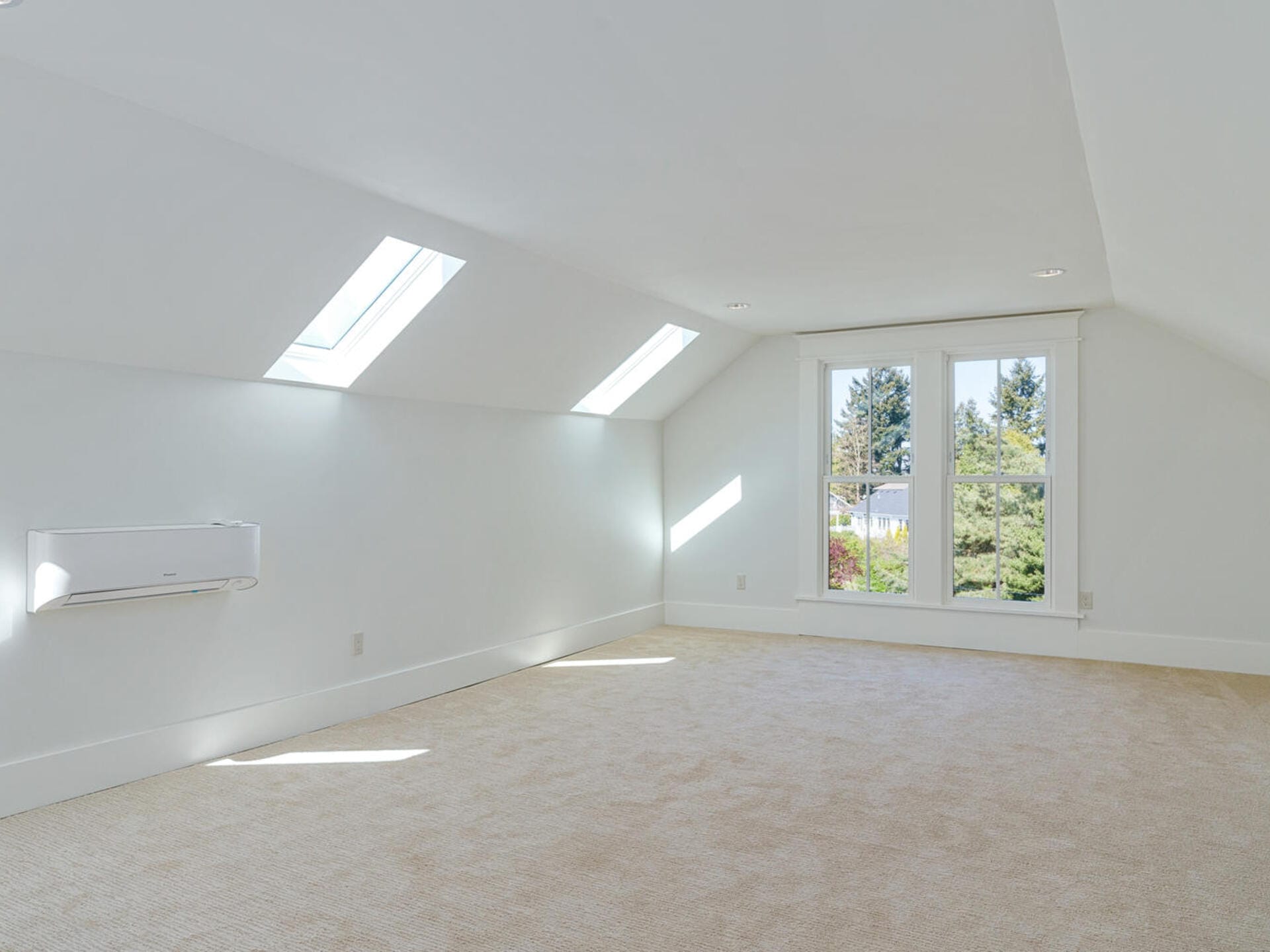 A bright, empty attic room with a neutral carpet, two skylights, and a large window offering a view of trees. The walls and ceiling are painted white, creating a clean and airy atmosphere.