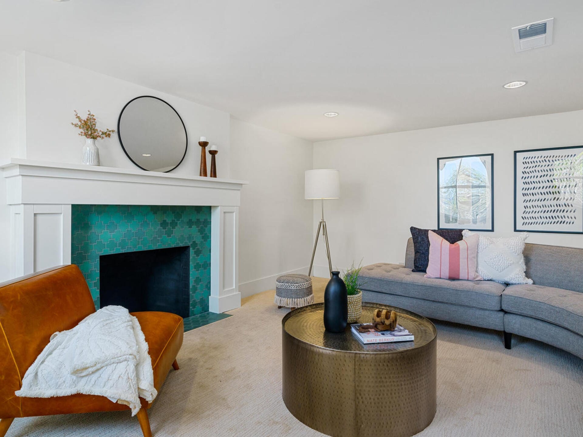 A modern living room with a teal-tiled fireplace, round mirror, and decorative vases on the mantel. A brown leather chair, gray sectional sofa with colorful pillows, and a round coffee table with magazines. A floor lamp and framed art complete the decor.