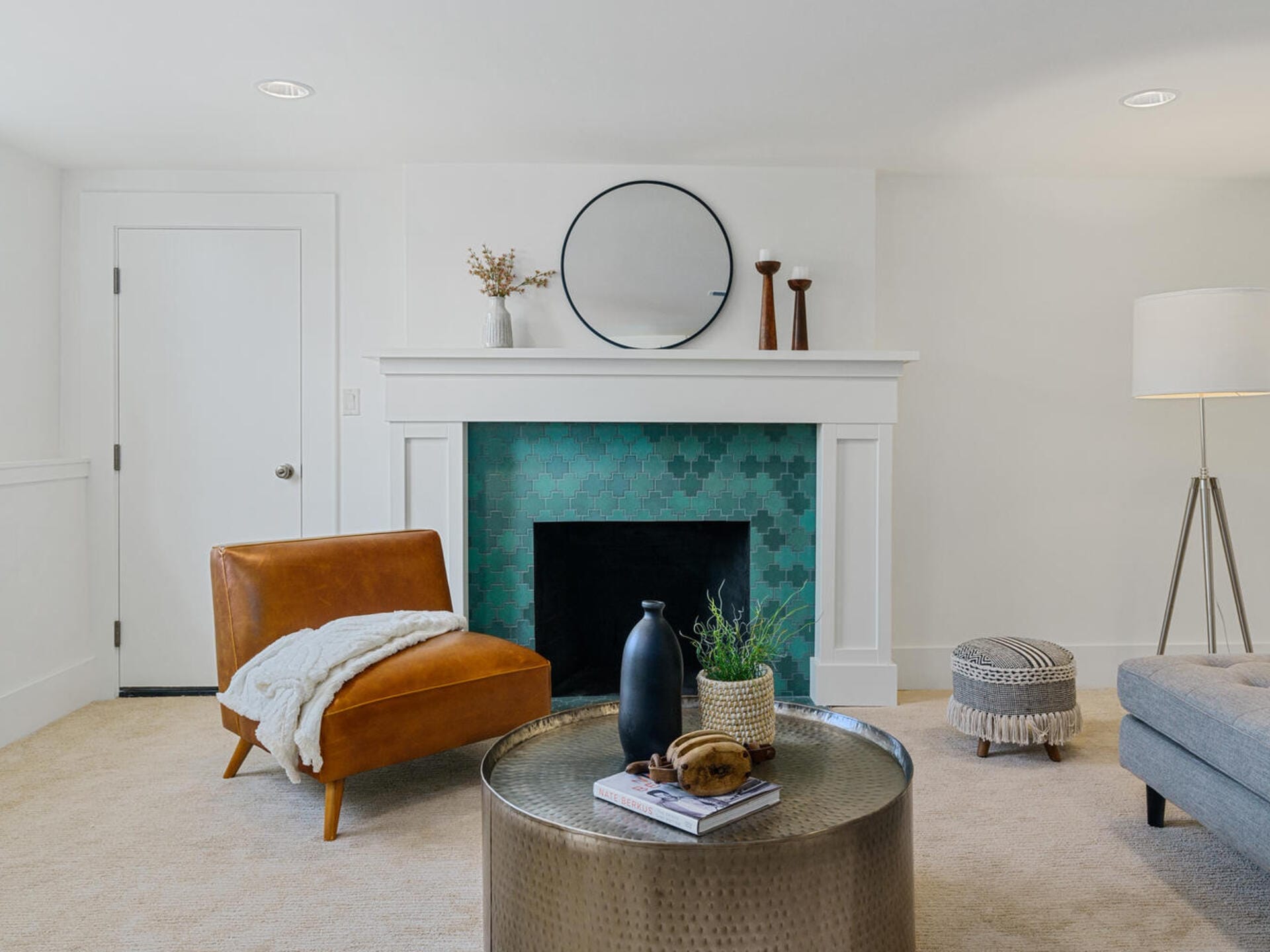 A modern living room featuring a fireplace with teal tile surround, a round mirror above it, and decorative items on the mantel. A brown leather chair with a white throw, a round metallic coffee table, and a gray pouf enhance the cozy atmosphere.