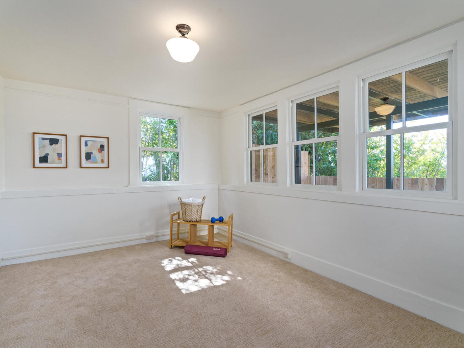 A bright, empty room with beige carpet, cream walls, and several windows. Two abstract paintings hang on a wall. In the corner, theres a small wooden chair, a basket, and two small dumbbells.