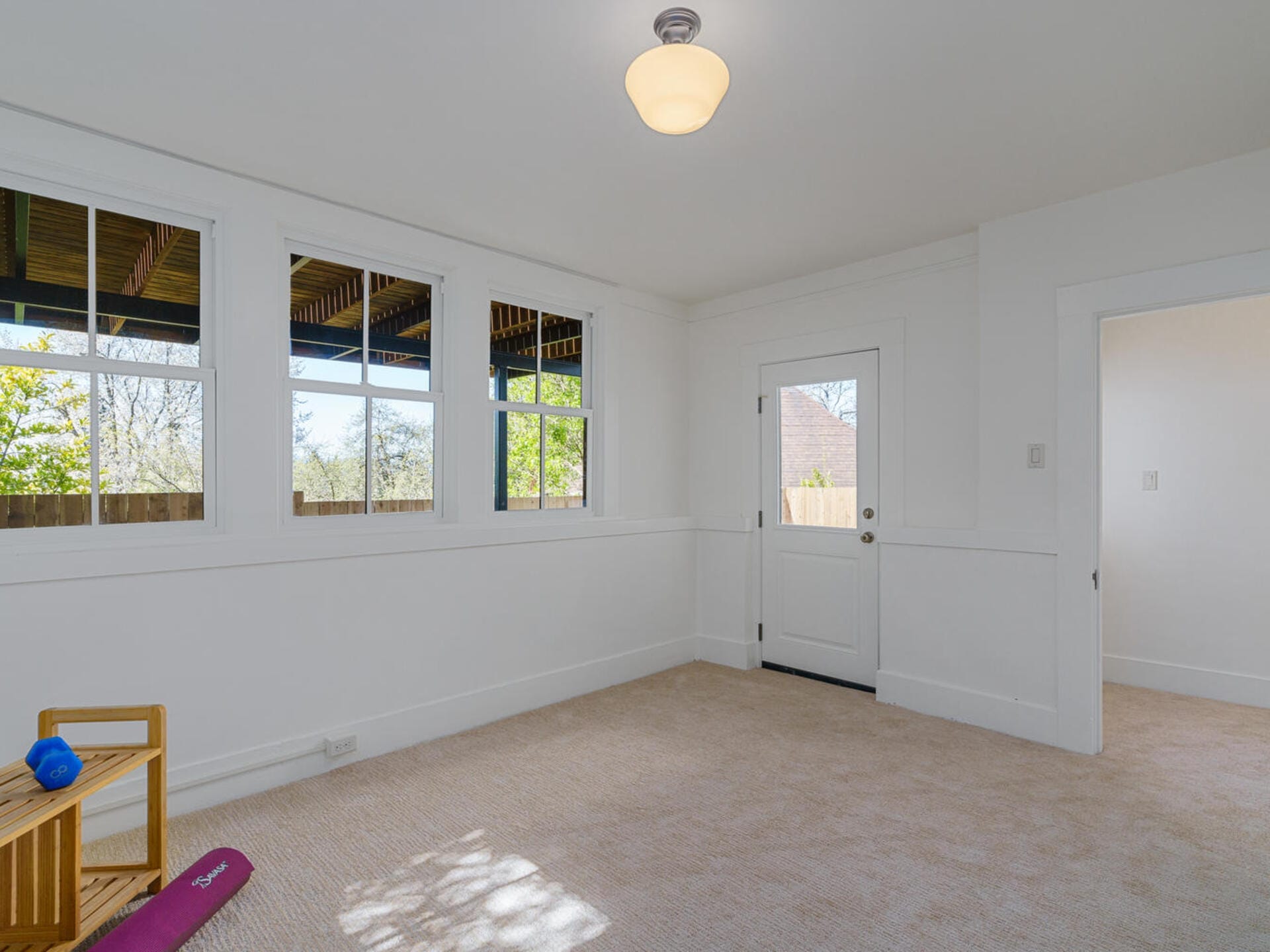 A bright, empty room with beige carpet, white walls, and a single ceiling light. Three large windows let in natural light. A closed door is on the right. A small wooden rack with a purple yoga mat and blue dumbbell is in the corner.