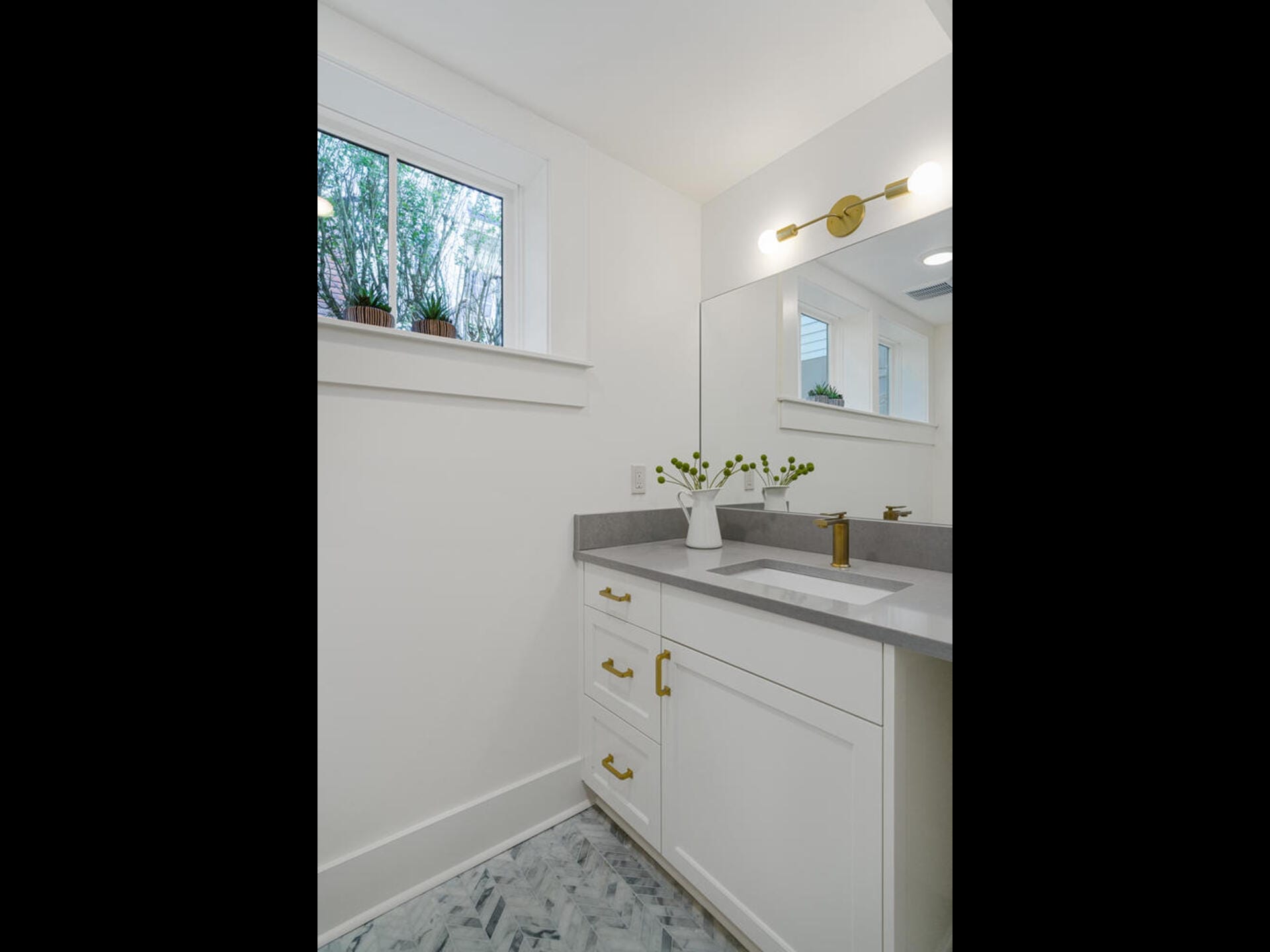 A modern bathroom with white walls and a marble floor. It features a white vanity with brass fixtures, a large mirror, and a gray countertop. A window with potted plants brings in natural light.