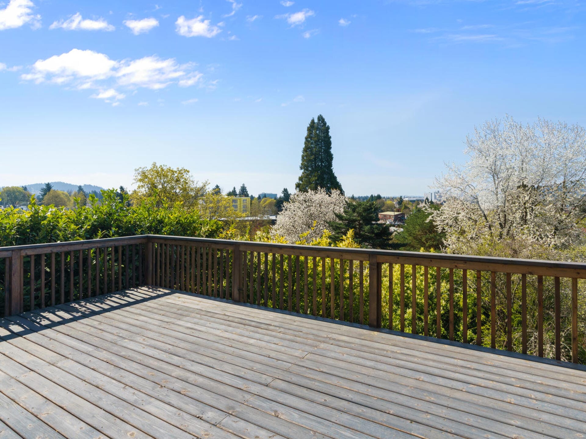 Wooden deck overlooking a scenic view of trees, including a large evergreen and blooming white trees, under a clear blue sky with a few clouds.