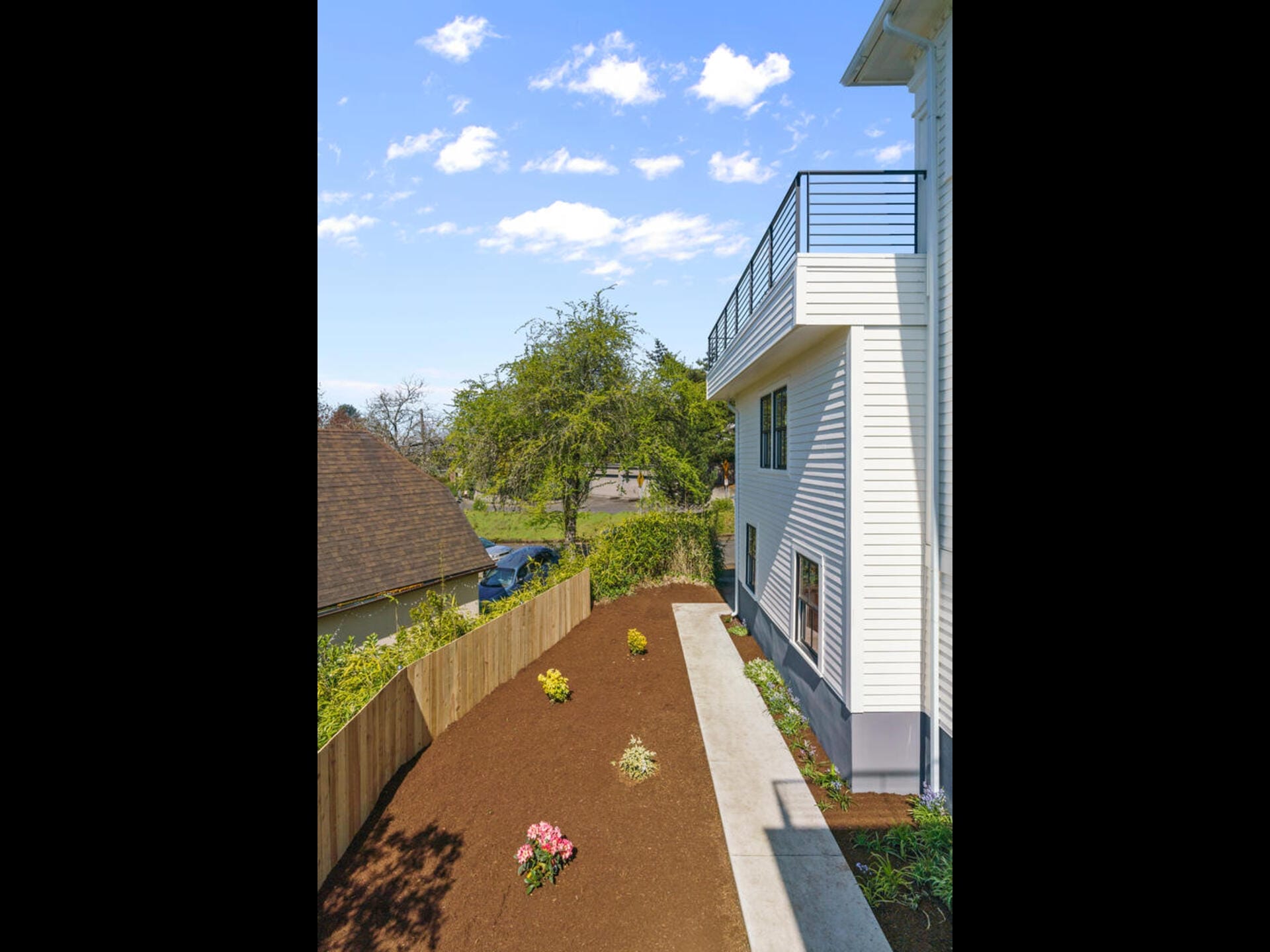 Side view of a modern house with white siding, featuring a balcony with metal railings. A newly landscaped garden with small plants and flowers is bordered by a wooden fence. The sky is clear with scattered clouds. A neighboring house is visible.