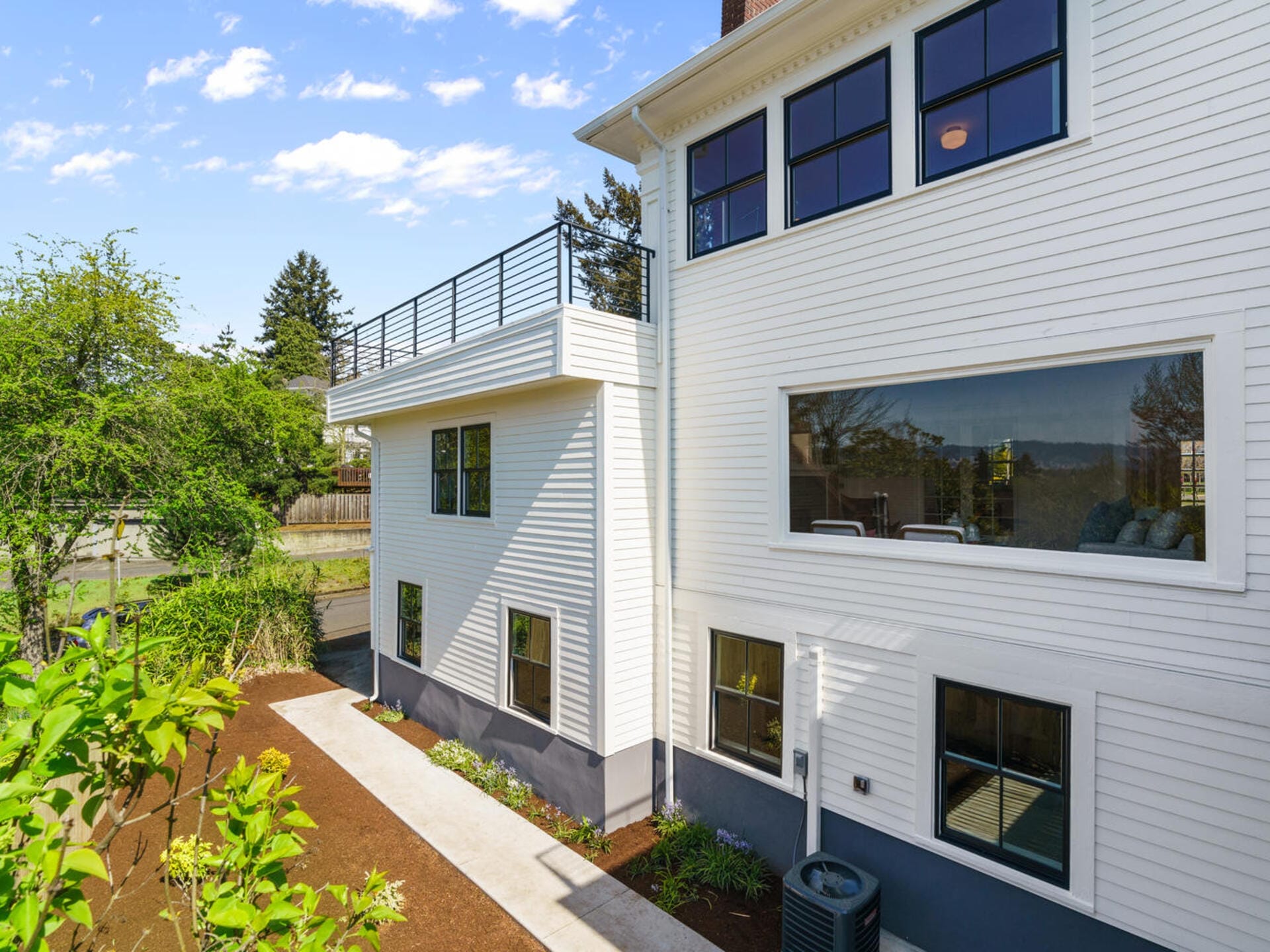 A modern white house with large windows and a rooftop terrace. The side yard features a concrete path next to a freshly planted garden. A blue sky with scattered clouds is visible above.