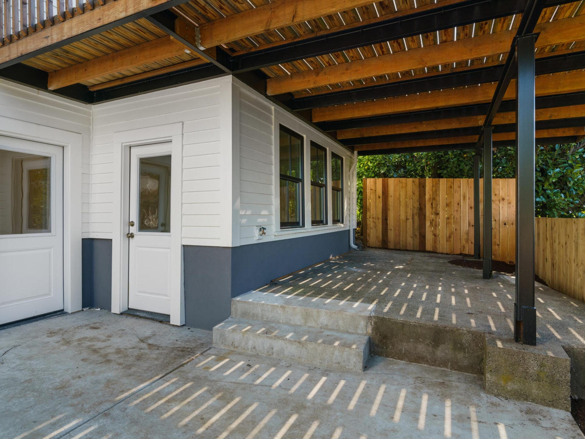 A covered outdoor area with wooden beams and slatted roof casting shadows on the ground. Two white doors, one with a frosted glass panel, lead into a building with white siding and large windows. A wooden fence is visible in the background.