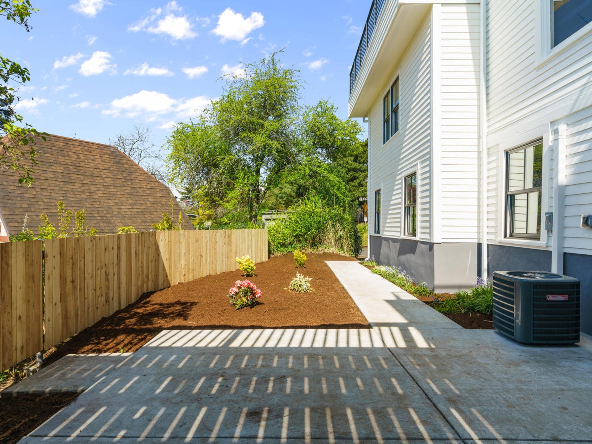 A tidy backyard shows a concrete path surrounded by mulched garden beds with small plants. A wooden fence lines one side, while the white exterior of a house forms the other boundary. An air conditioning unit is visible near the house.
