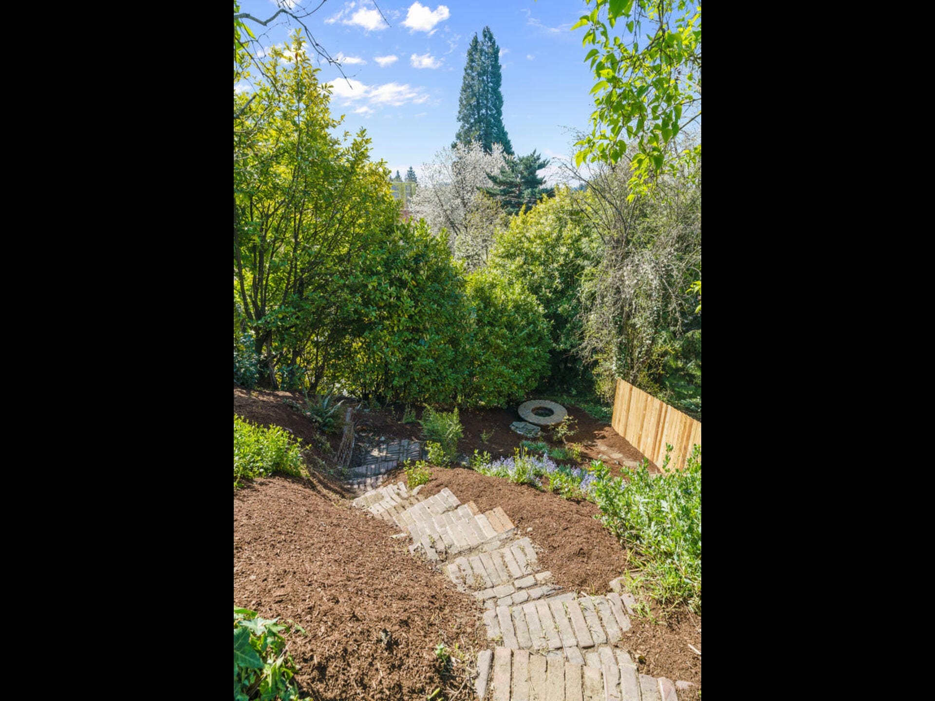 A stone path curves down a lush garden hill, bordered by green shrubs and trees. In the background, a wooden fence and a round stone structure are visible, with a tall evergreen tree and blue sky overhead.