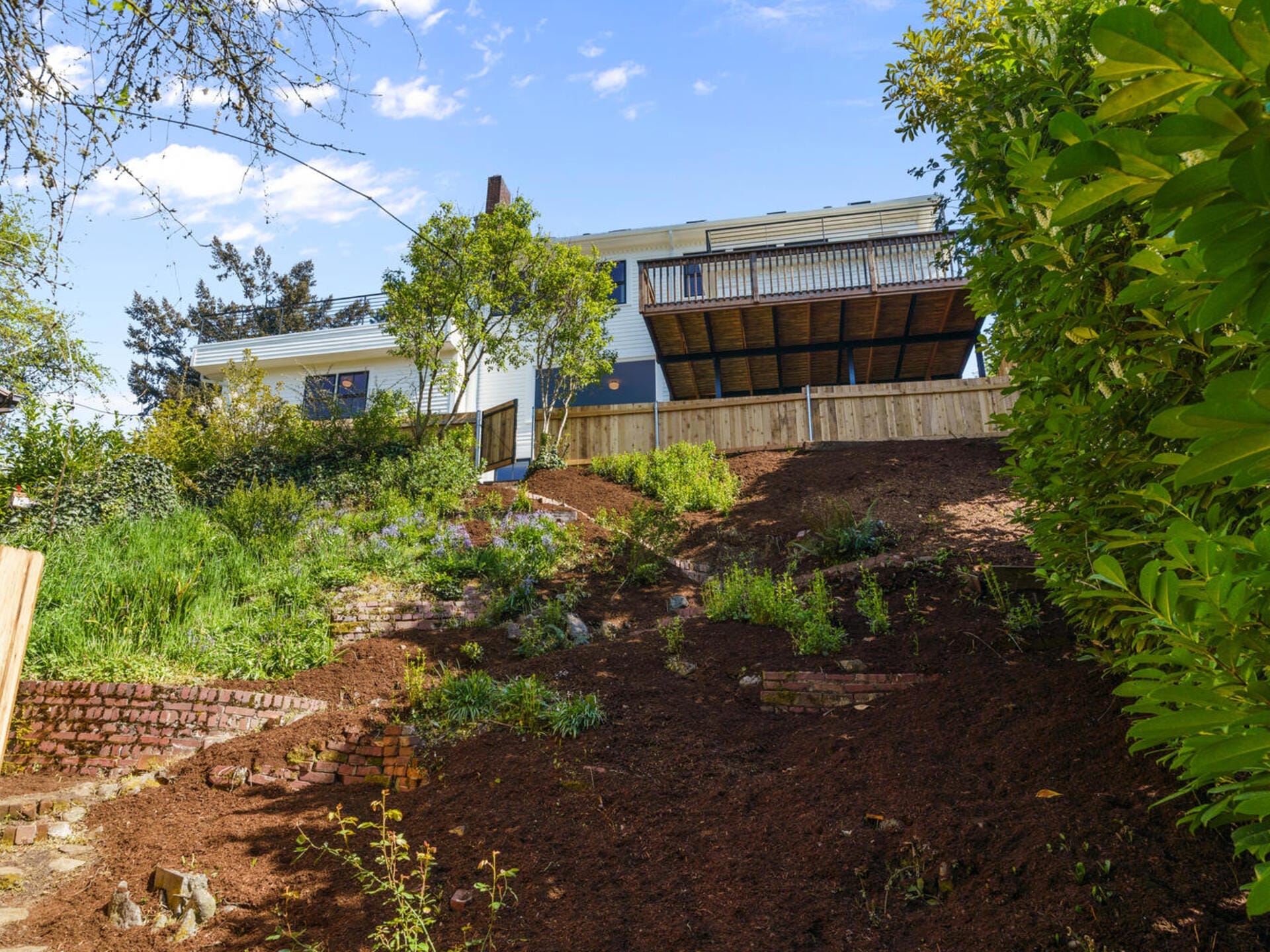 A house with a large deck overlooks a steeply sloped backyard with terraced garden beds. The area is partially covered in greenery and fresh soil, surrounded by trees and a wooden fence under a clear blue sky.