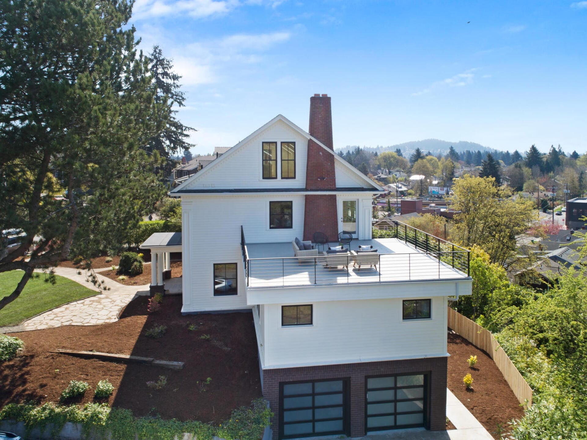 Aerial view of a large white house with a flat rooftop patio, surrounded by greenery. The house has multiple stories with large windows and a red brick chimney. A spacious driveway leads to two garage doors. Scenic background with trees and hills.