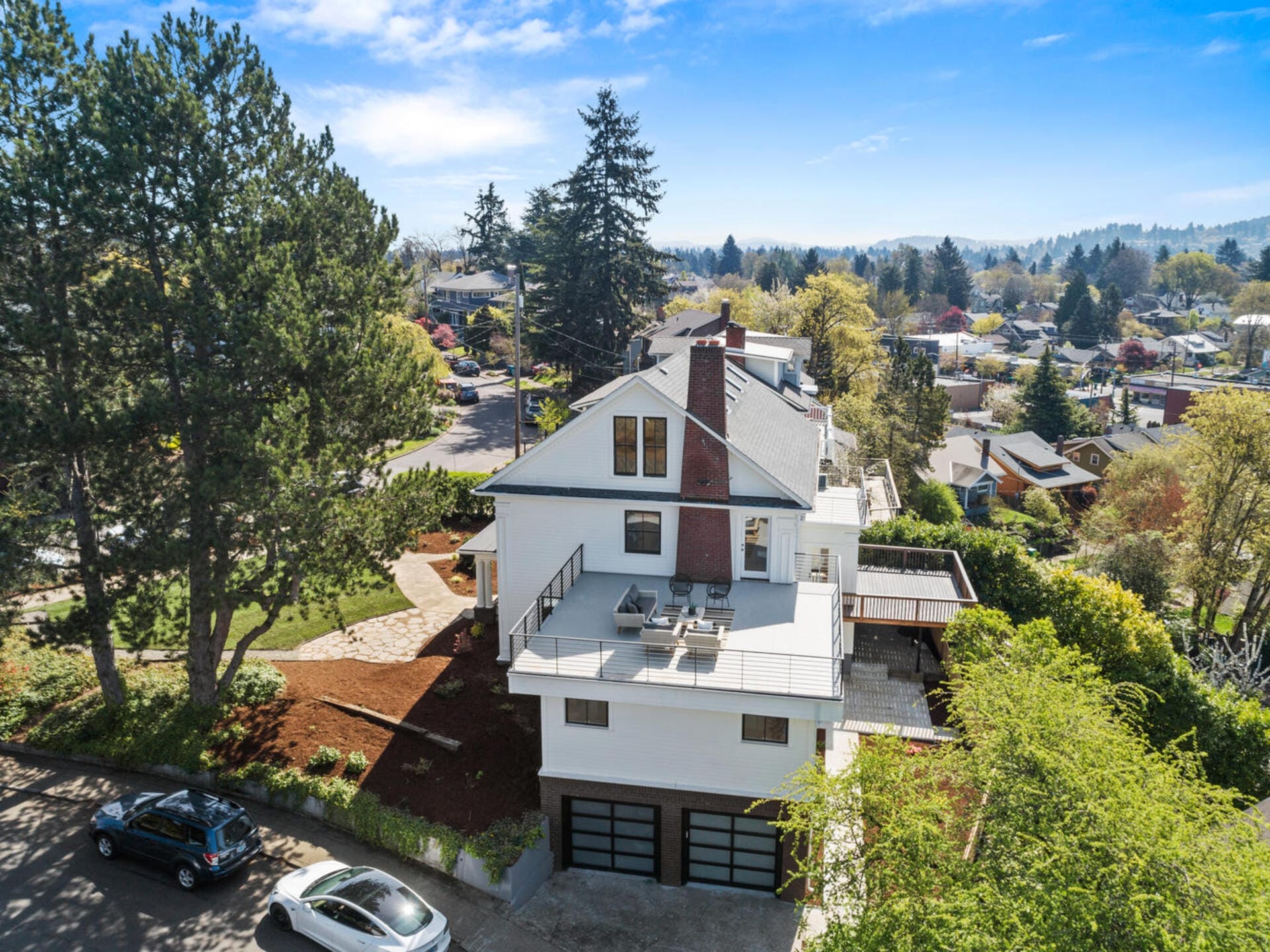 Aerial view of a large white house with a rooftop patio and multiple balconies. The house has a sloped roof and is surrounded by trees and a well-maintained garden. A car is parked on the driveway. The neighborhood features similar homes and lush greenery.