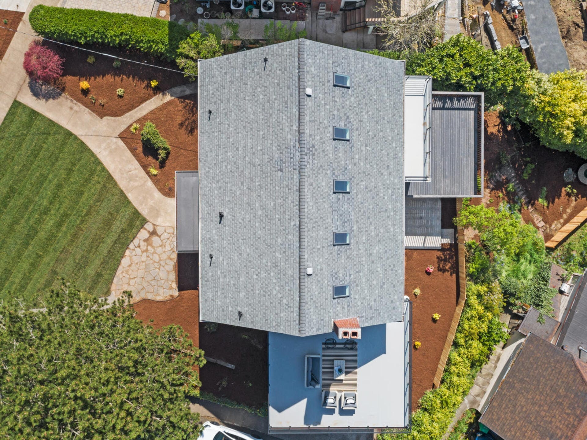 Aerial view of a modern house with a gray roof, surrounded by a garden and landscaped yard. The garden has a mix of grass, pathways, and various plants. Theres a paved driveway and several trees in the vicinity.