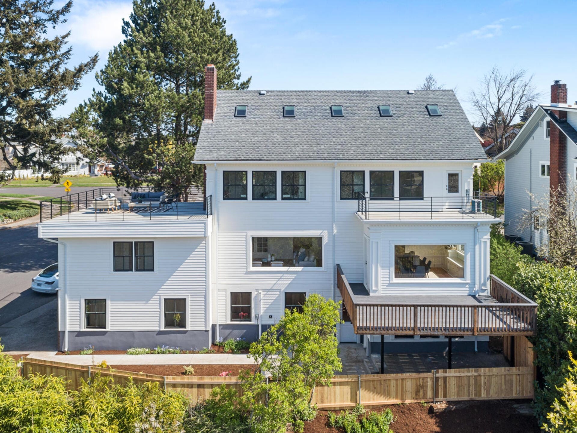 A large, modern white house with multiple stories, skylights, and a spacious wooden deck. Surrounded by trees and a wooden fence, it stands on a corner lot under a clear blue sky.