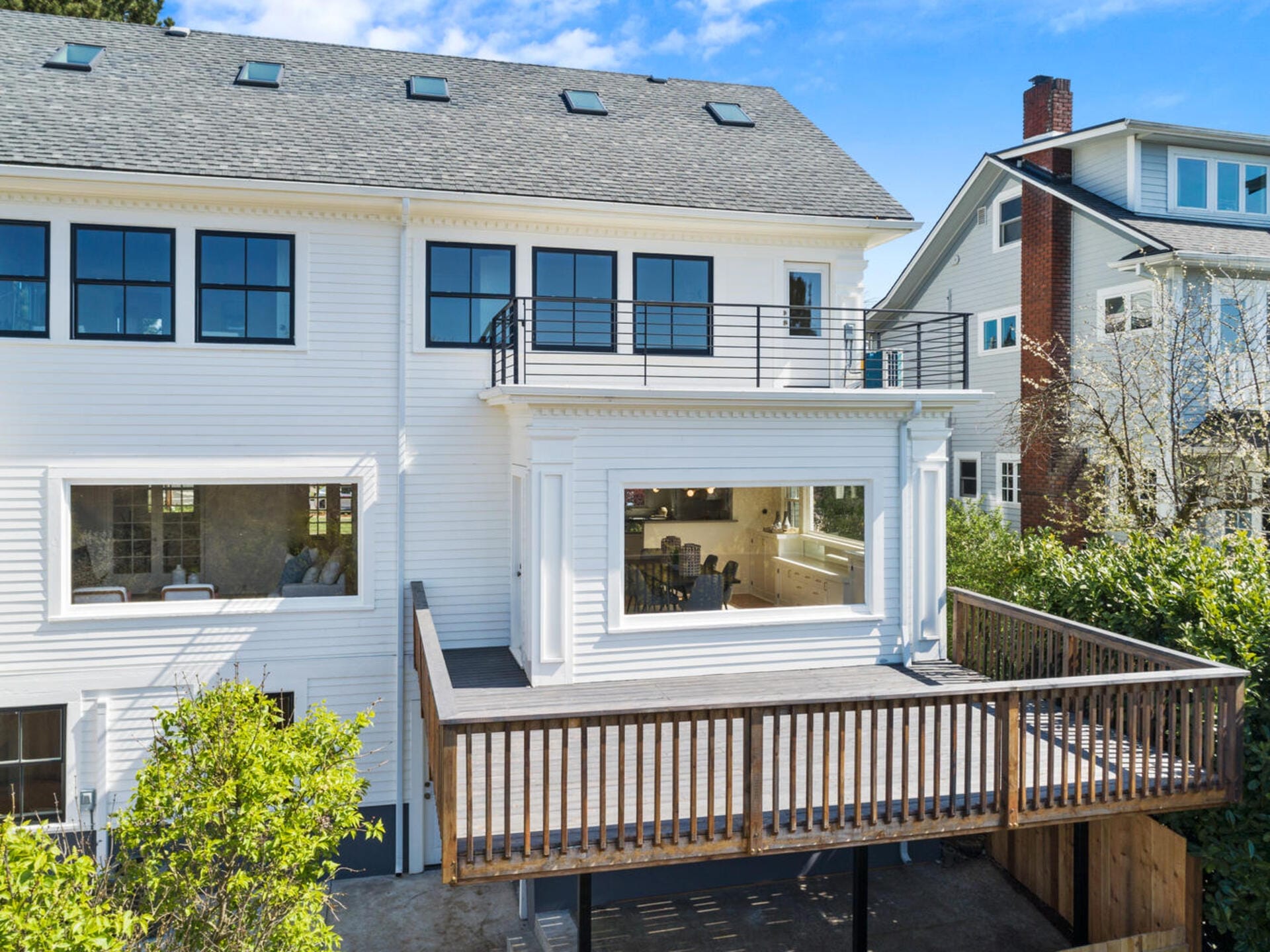 A two-story white house with large windows and a gray roof. It features a wooden deck extending from the second floor. The house has a modern design with a spacious balcony, surrounded by greenery and neighboring homes.