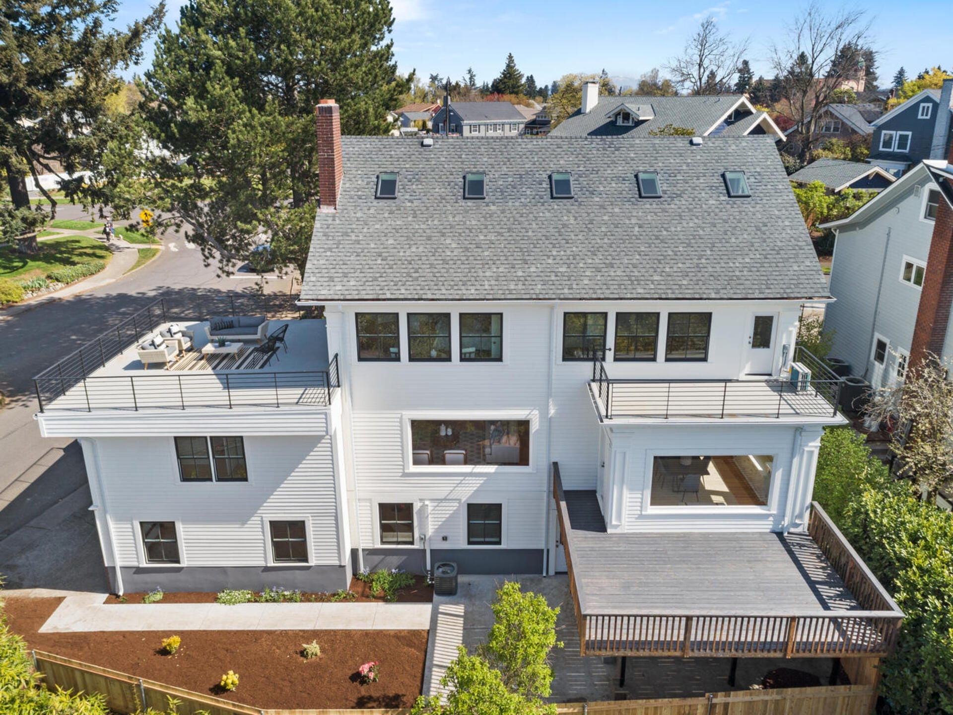 A large white multi-story house with multiple windows and a balcony. The roof has several skylights, and theres a spacious wooden deck on the side. The house is surrounded by trees and additional homes in the background.