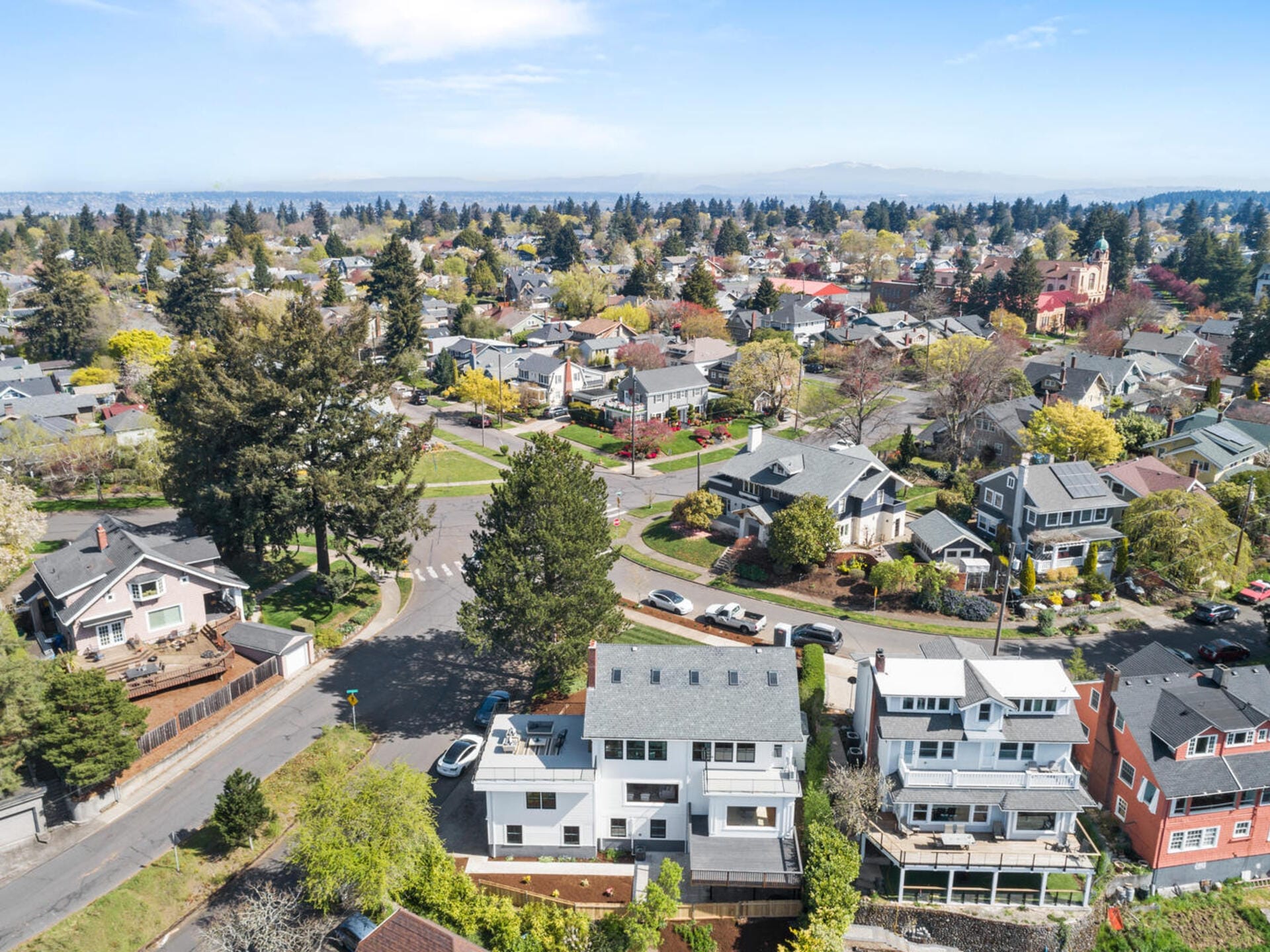 Aerial view of a suburban neighborhood with a mix of houses, trees, and streets. The area is lush with greenery and several residential homes are visible. The sky is clear, indicating a sunny day.
